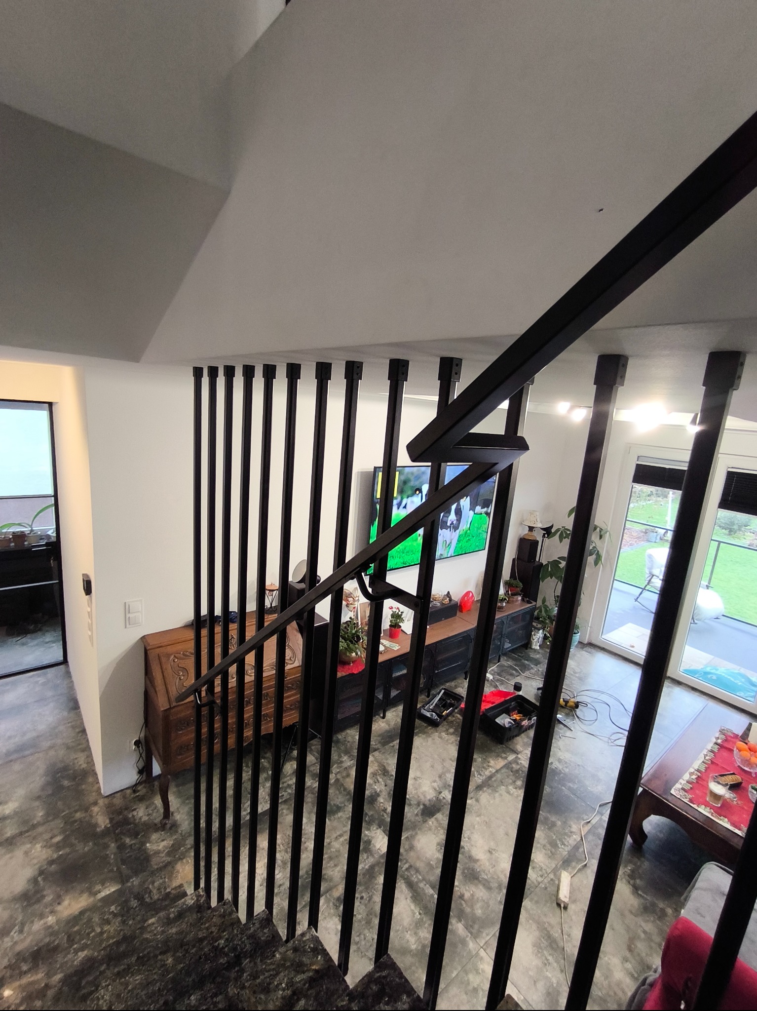 Nowoczesne schody z czarną metalową balustradą, widok z góry na salon z telewizorem i meblami, na podłodze widoczne narzędzia i przedłużacz.