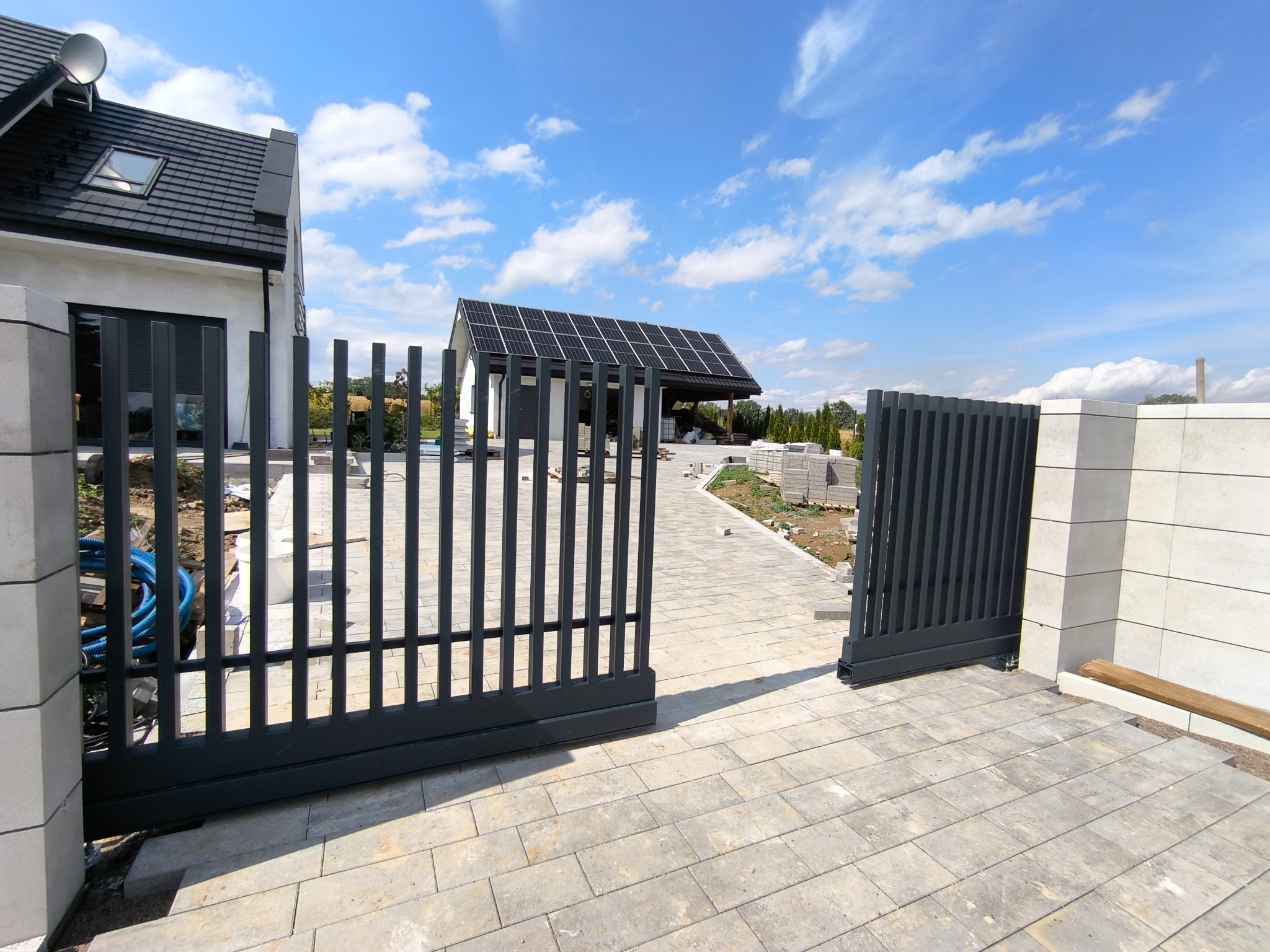 Nowoczesna, grafitowa brama wjazdowa, z pionowymi szczeblami, na tle jasnej kostki brukowej i domu z panelami słonecznymi. Brama otwarta, widok z zewnątrz.