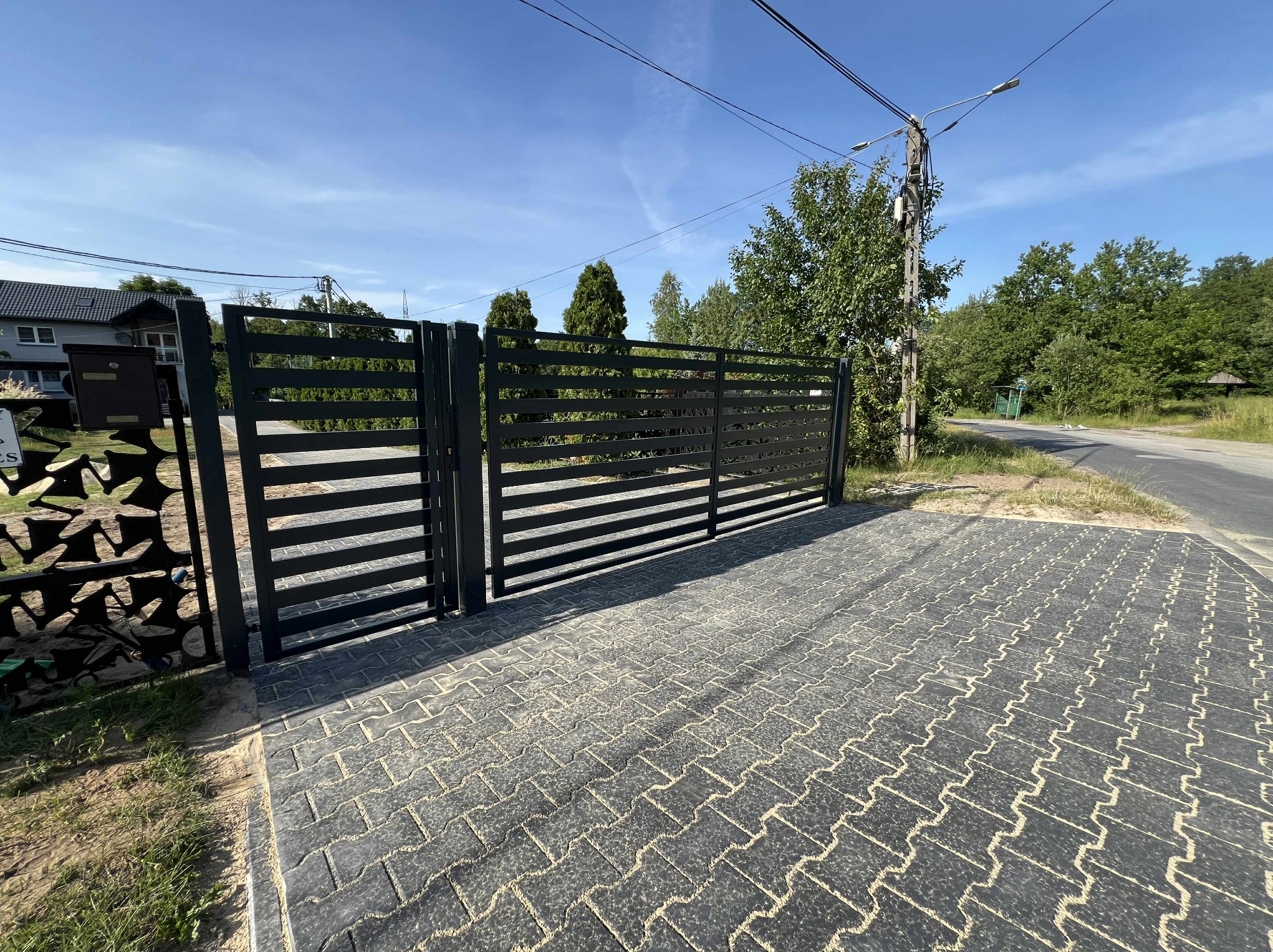 Nowoczesna, pozioma brama wjazdowa w kolorze antracytu na tle brukowanej posesji i zieleni. Widoczny fragment furtki z dekoracyjnym motywem. Słoneczny dzień.