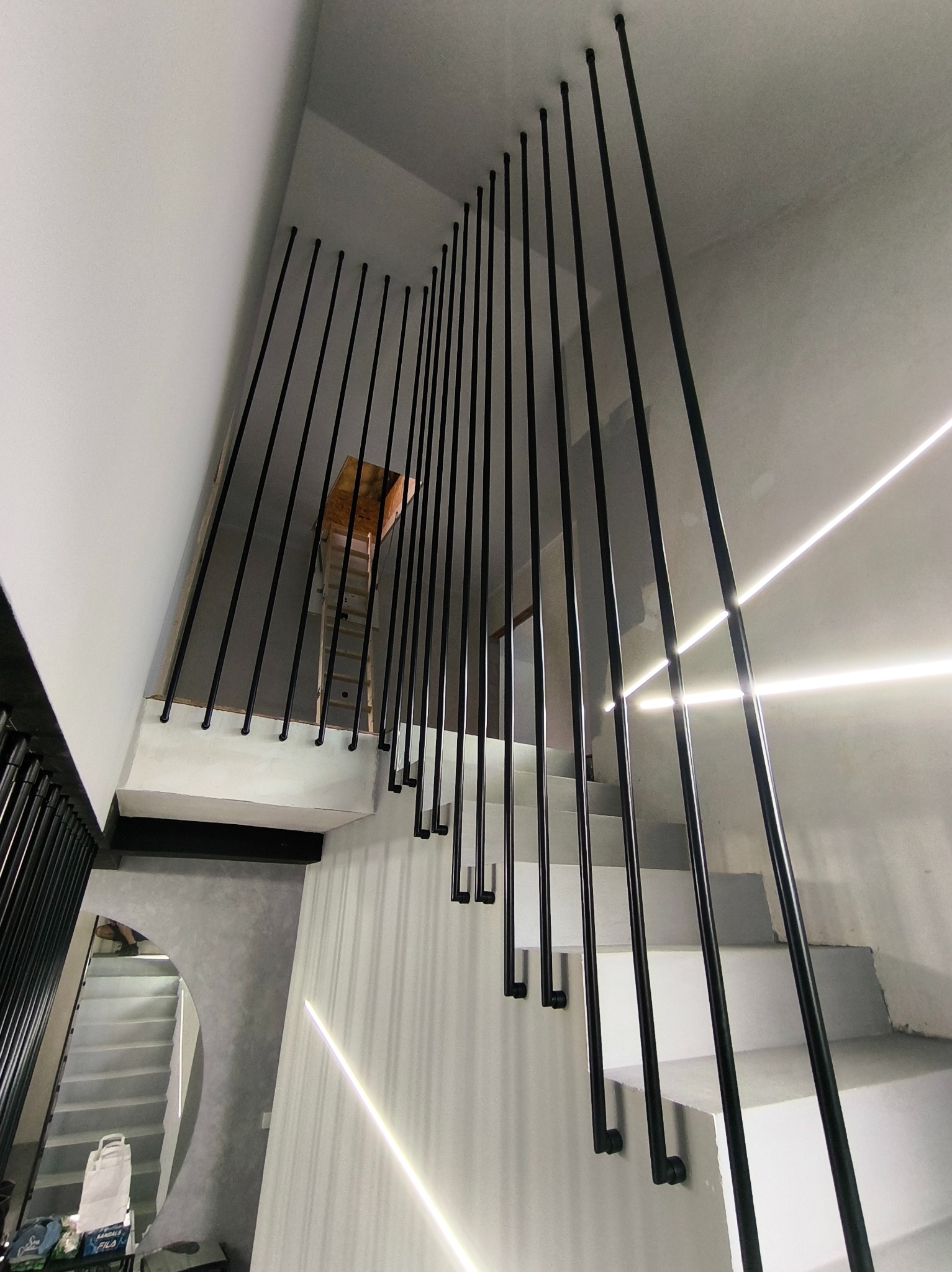 Nowoczesne schody z czarnymi, pionowymi balustradami, oświetlone listwami LED, widziane z dołu. W tle drabina i fragment lustra. Minimalistyczny design.