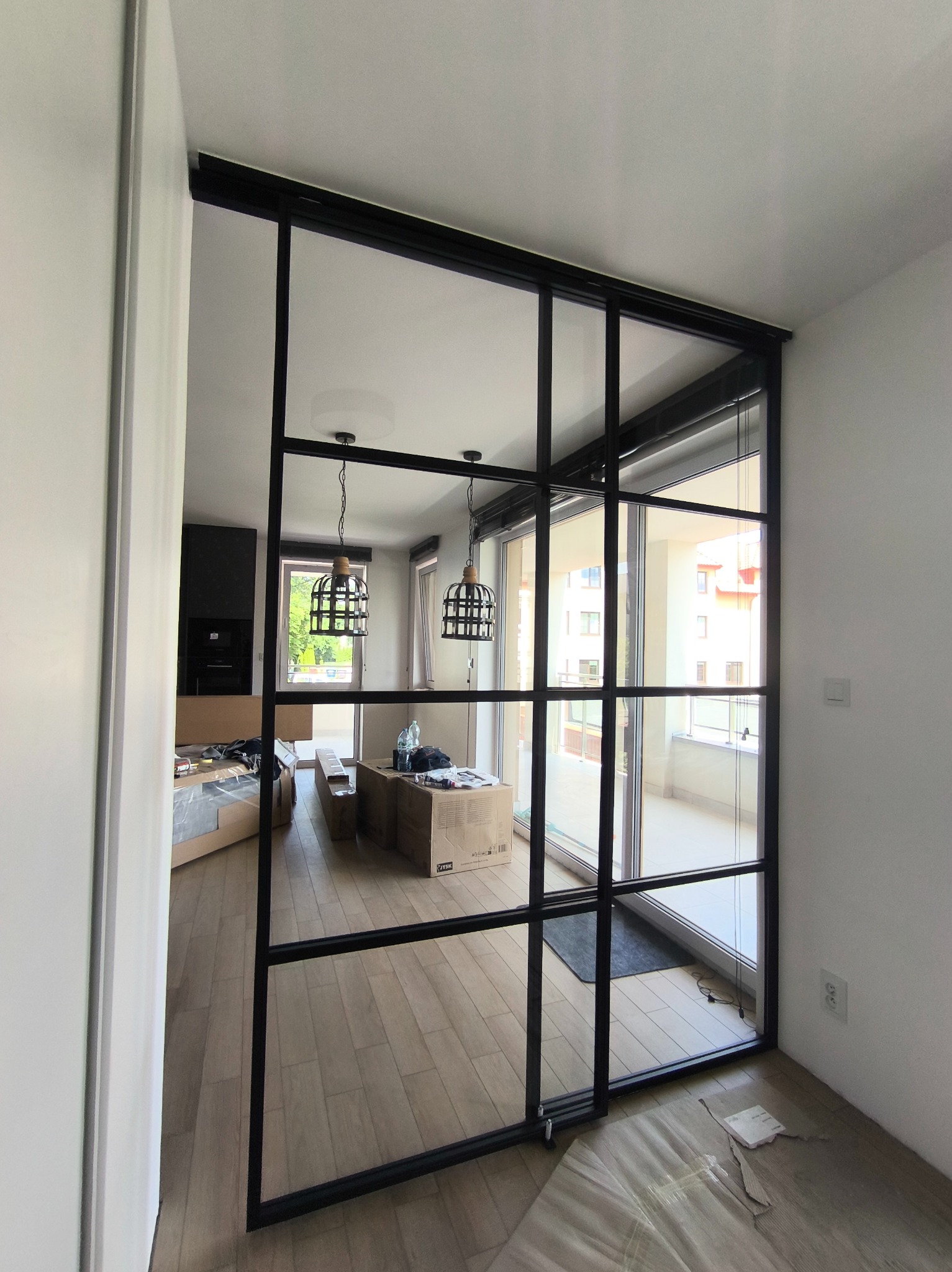 Nowoczesna, czarna, metalowa konstrukcja z przeszkleniami, tworząca ściankę działową między pokojem a balkonem. Widoczne elementy montażowe i częściowo rozpakowane kartony w tle.