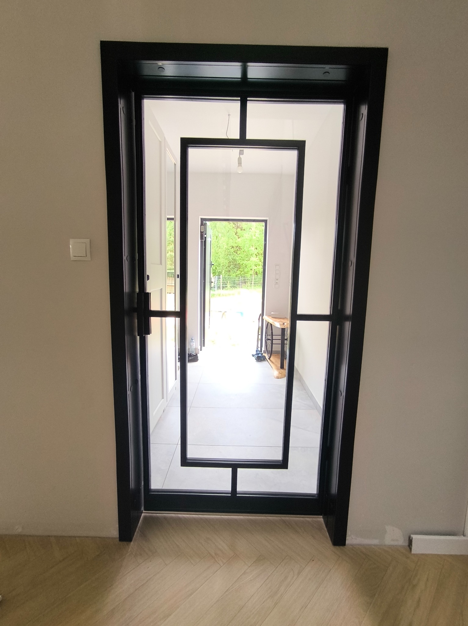 Nowoczesne, czarne drzwi metalowe z geometrycznym wzorem, widok z wnętrza domu na zewnątrz. Jasne wnętrze, drewniana podłoga, minimalistyczny design.