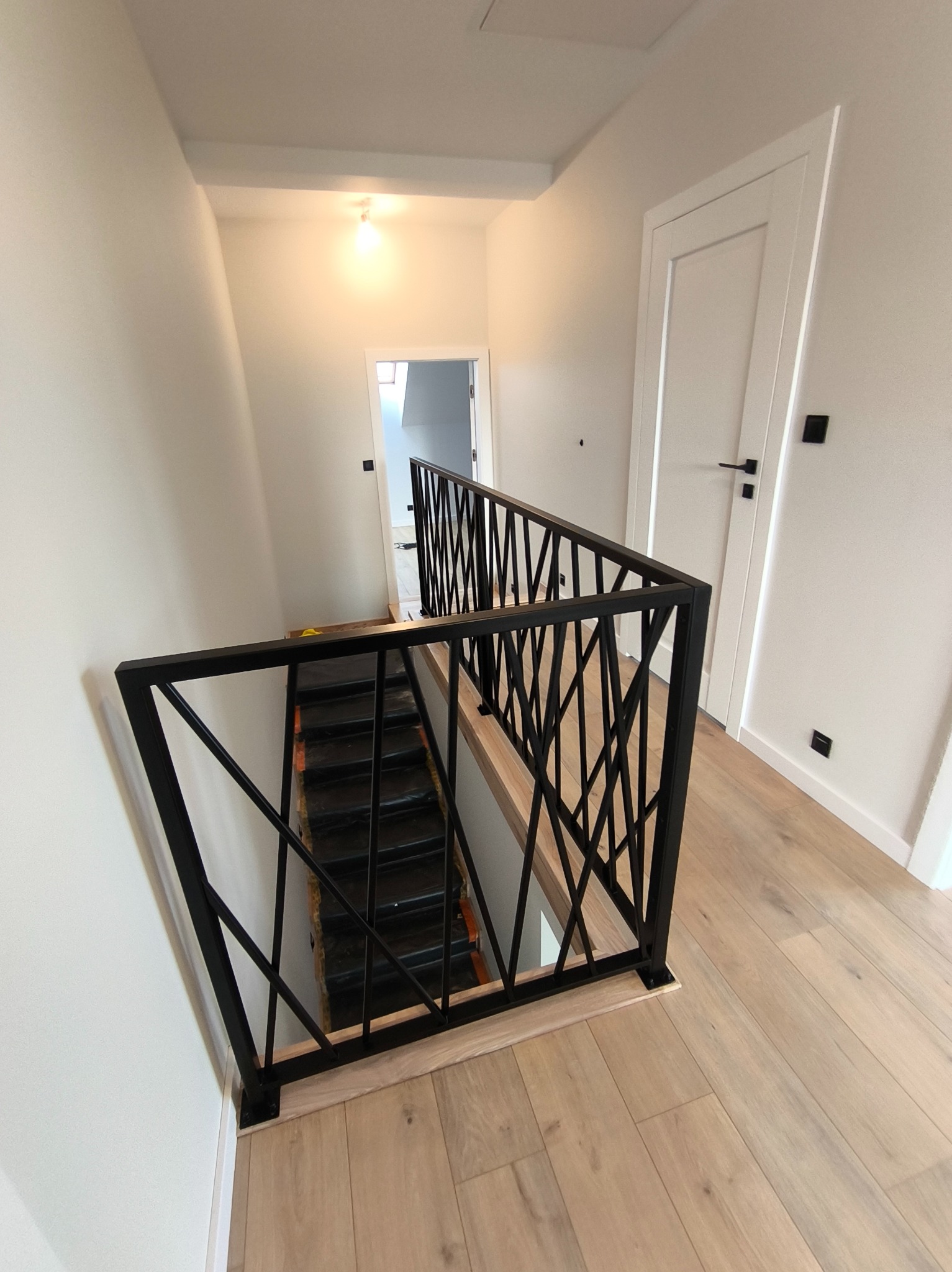 Nowoczesna balustrada metalowa w kolorze czarnym z geometrycznym wzorem, zamontowana na schodach w jasnym wnętrzu. Widok z góry na dół, parkiet na podłodze.