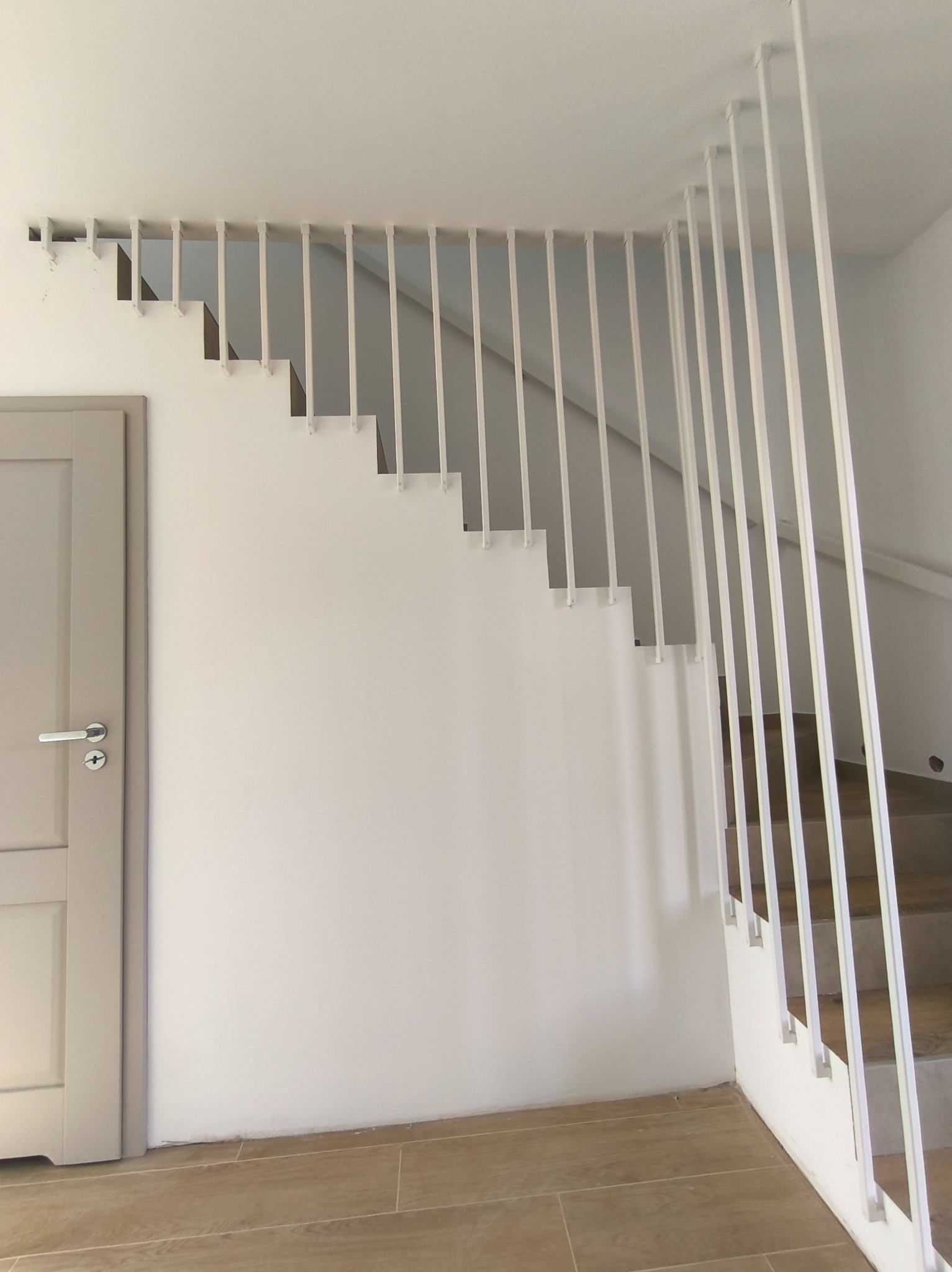 Minimalistyczne schody z białą, pionową balustradą, widoczne z boku. Szara ściana i drzwi po lewej stronie, drewnopodobna podłoga. Nowoczesny design.