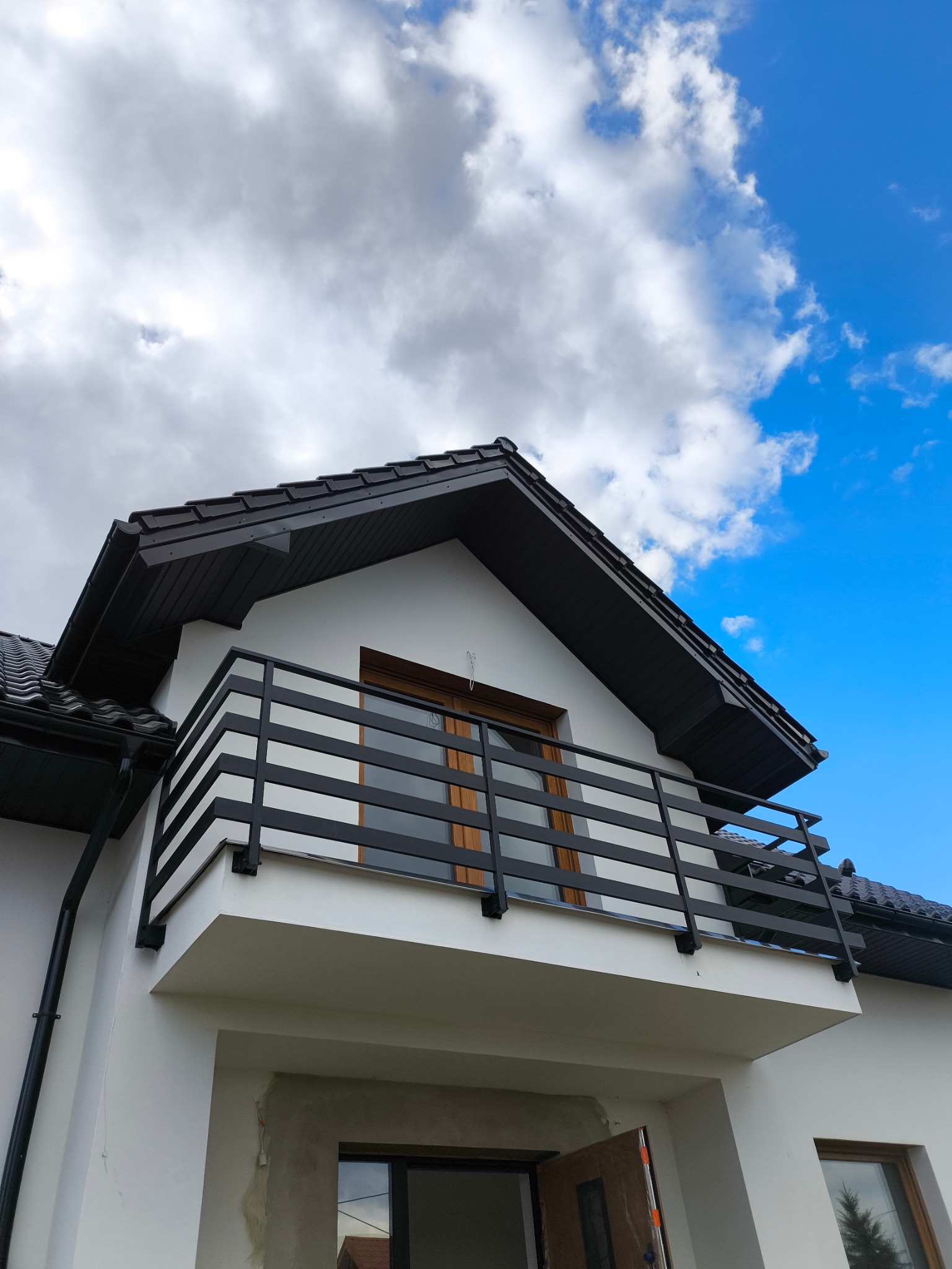 Nowoczesny balkon z czarną, metalową balustradą o poziomym układzie szczebli, na tle błękitnego nieba z obłokami, widok z dołu, fragment elewacji budynku.