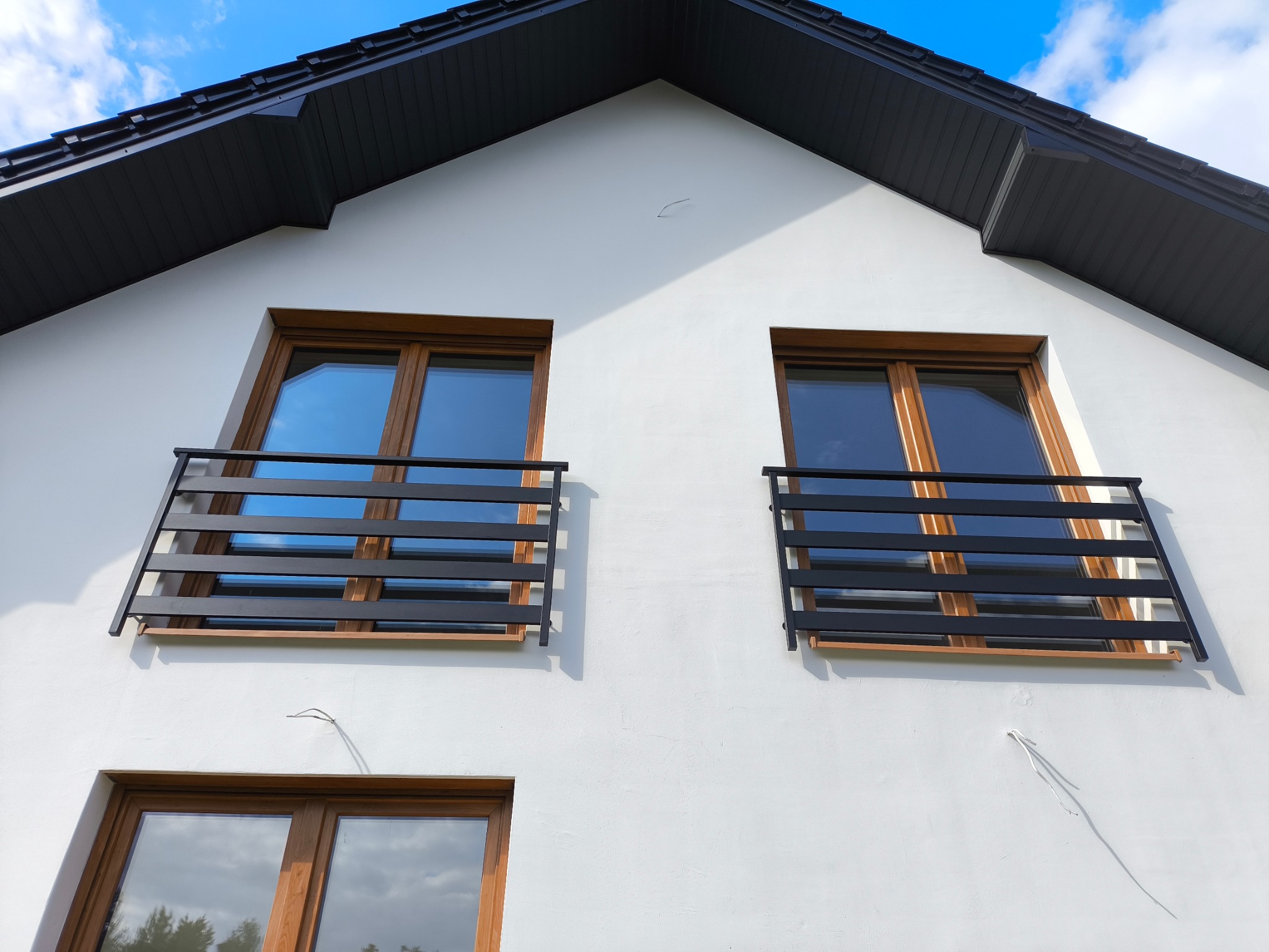 Elewacja domu z dwoma oknami balkonowymi zabezpieczonymi czarnymi, poziomymi balustradami. Widoczne brązowe ramy okienne i fragment dachu.