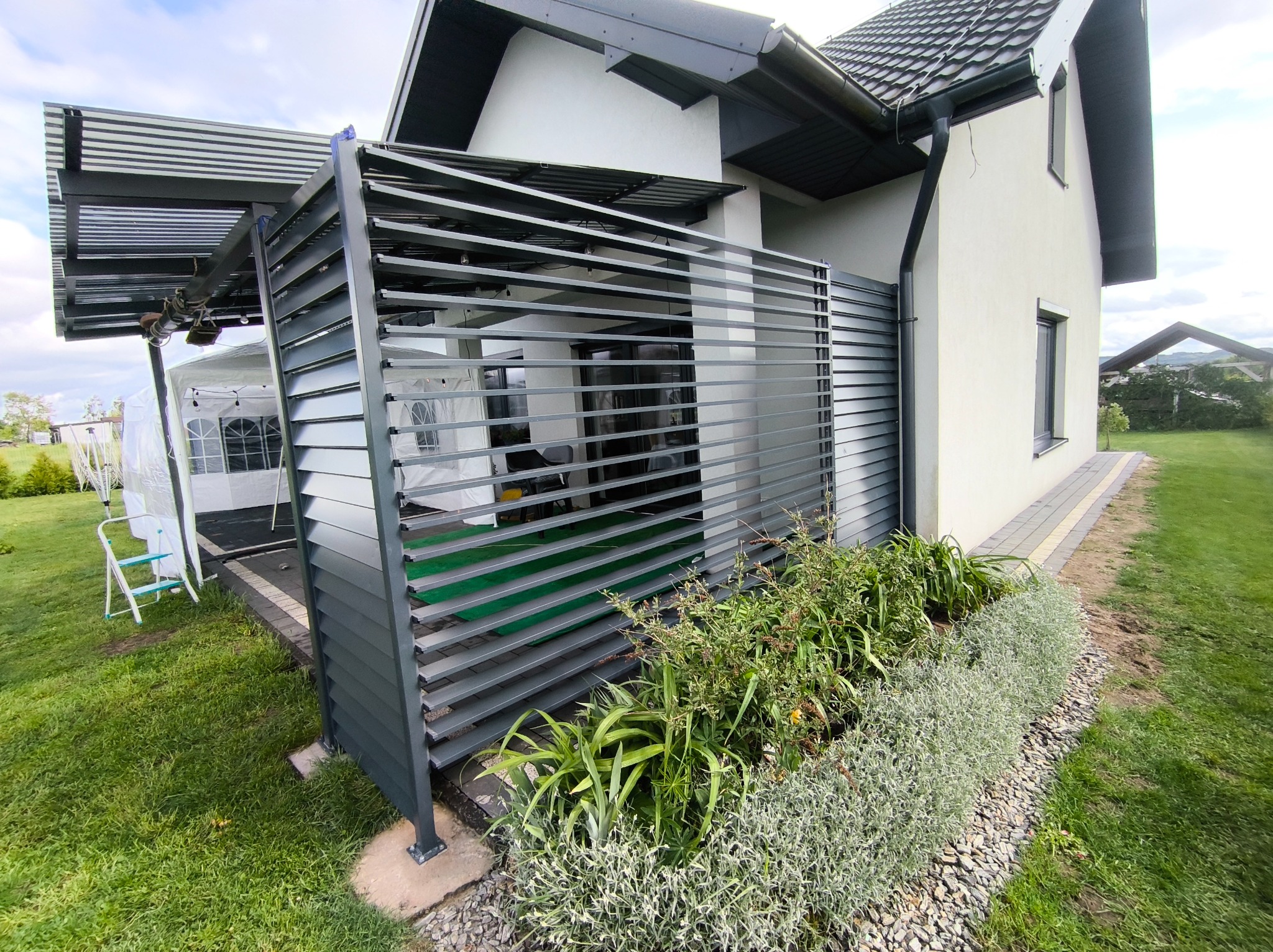 Nowoczesna pergola z szarymi, poziomymi lamelami przylegająca do białego domu. Widoczne rośliny wzdłuż konstrukcji i biały namiot w tle. Całość w zielonym ogrodzie.