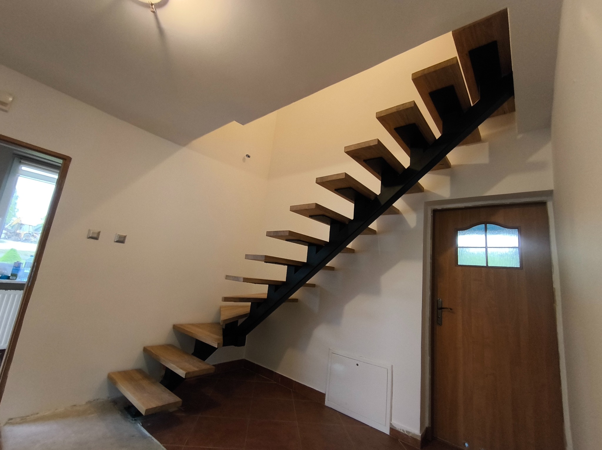 Nowoczesne schody metalowe z drewnianymi stopniami, wsparte na czarnej konstrukcji, w jasnym wnętrzu z brązową podłogą i drewnianymi drzwiami.