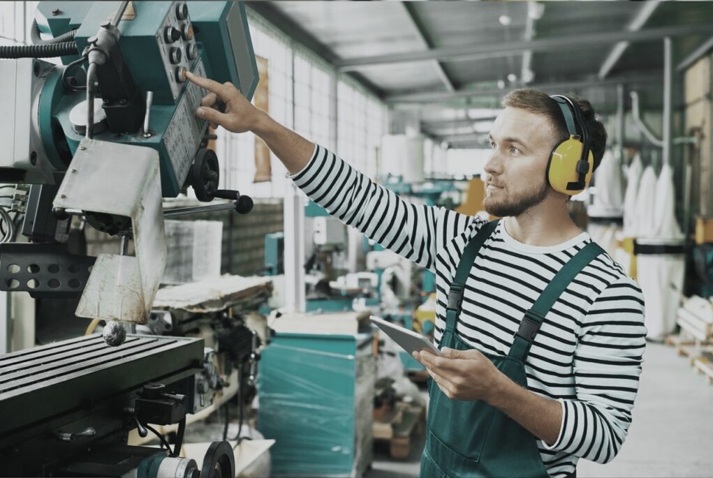 Pracownik w słuchawkach obsługuje maszynę przemysłową, trzymając tablet. Ubrany w kombinezon i bluzę w paski, w tle hala produkcyjna z maszynami.