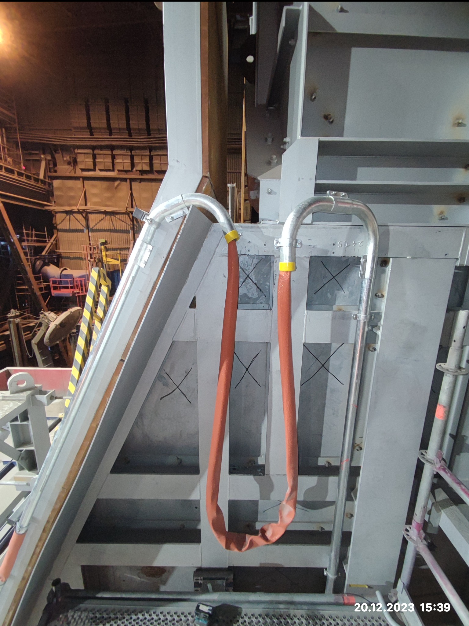 Szare metalowe elementy konstrukcyjne z rurami i pomarańczowymi osłonami, widoczne rusztowania i elementy instalacji w tle.