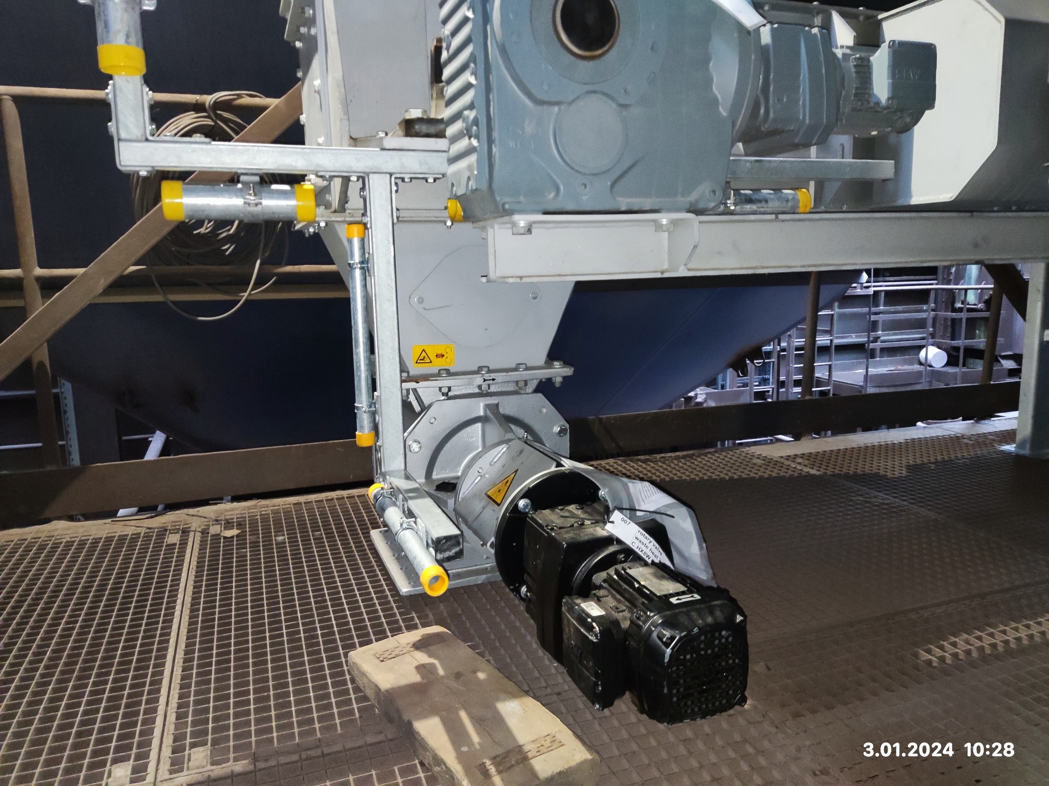 Zamontowany silnik elektryczny z przekładnią na metalowej konstrukcji w hali przemysłowej, widoczna tabliczka znamionowa i żółte elementy zabezpieczające.