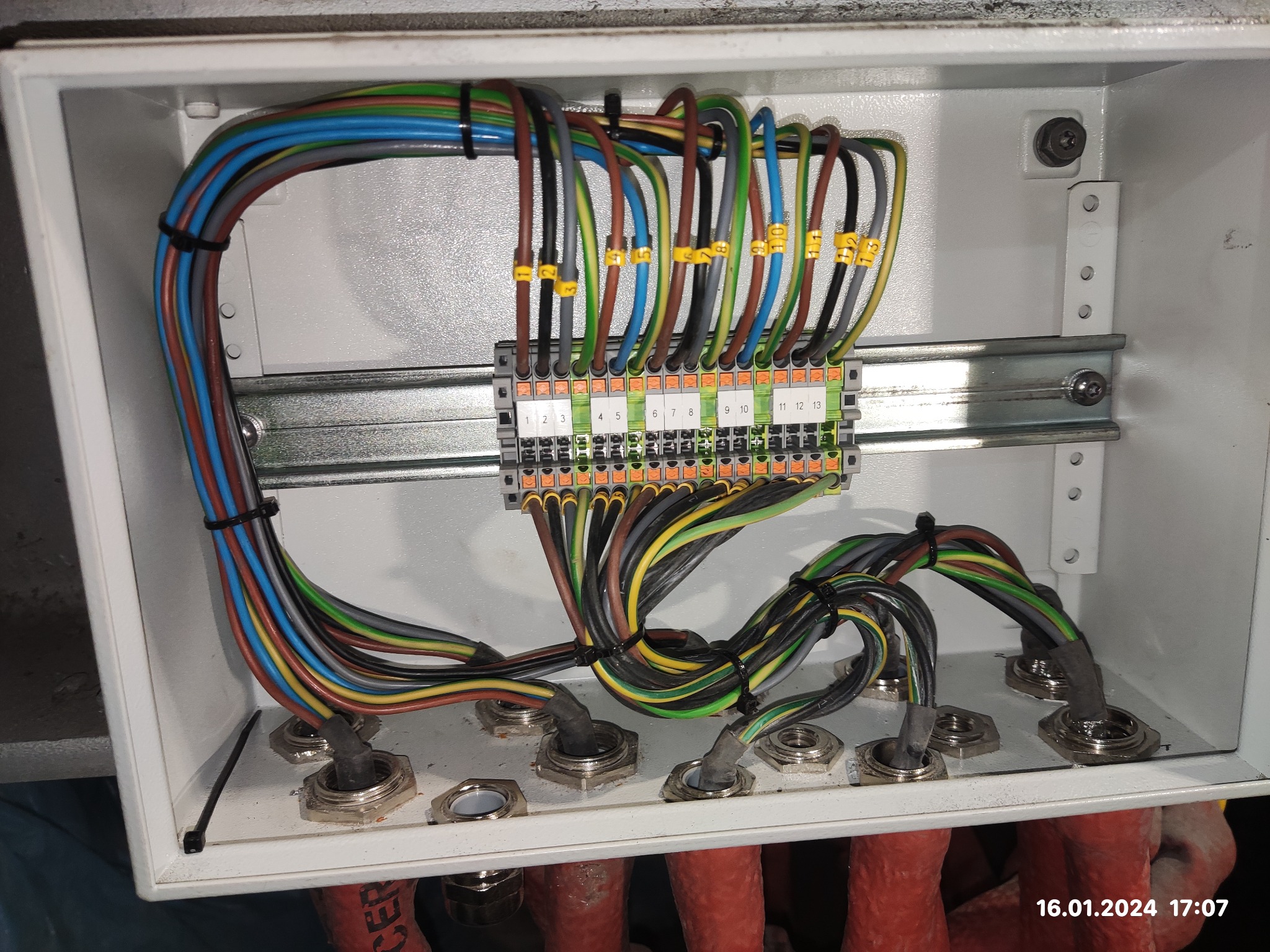 Otwarta skrzynka elektryczna z podłączonymi, ponumerowanymi przewodami w różnych kolorach, zabezpieczonymi czarnymi opaskami zaciskowymi, widoczne metalowe dławiki kablowe.