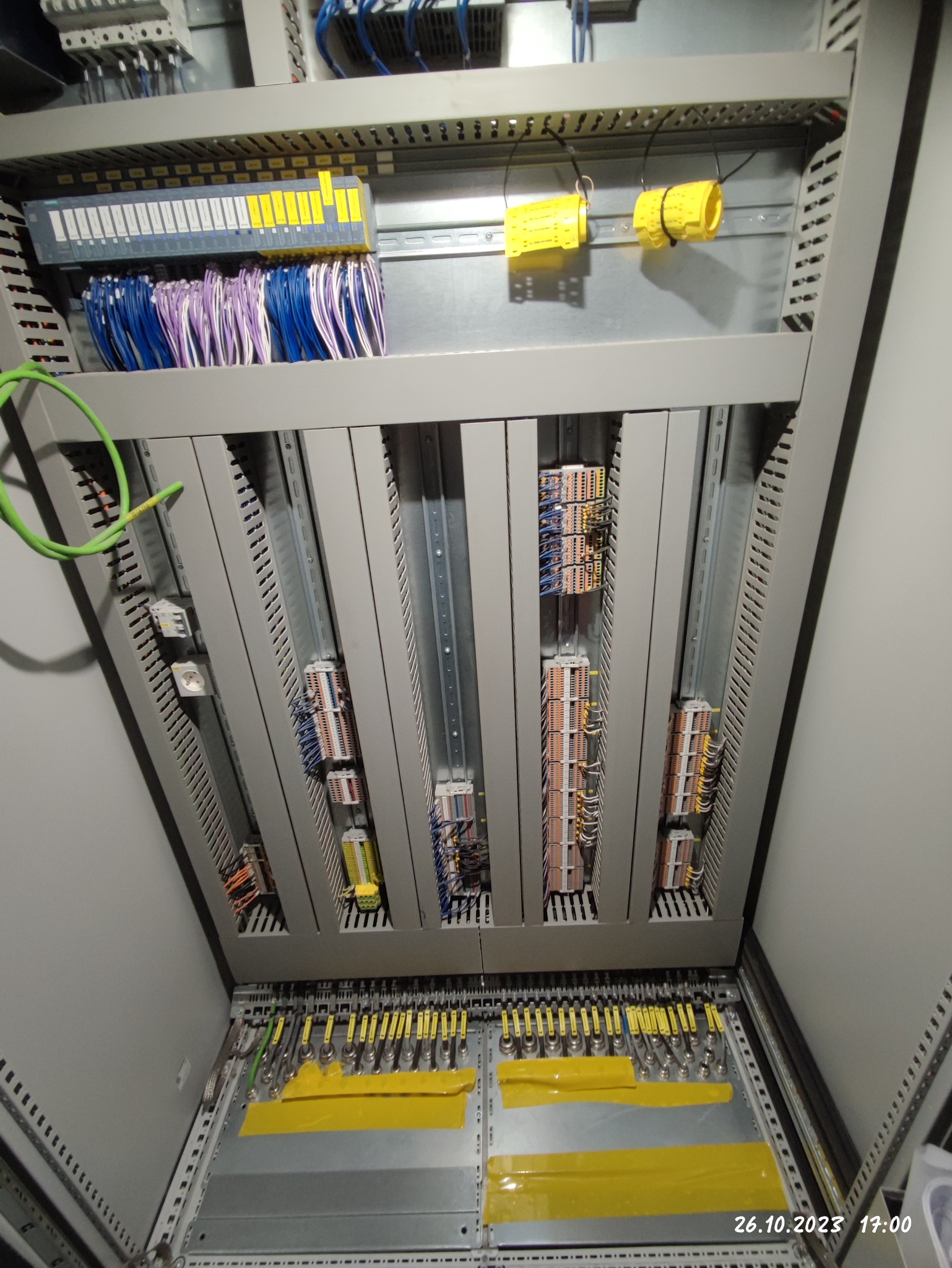 Wnętrze rozdzielnicy elektrycznej z modułami sterującymi, licznymi przewodami w różnych kolorach i żółtymi oznaczeniami na listwach zaciskowych.