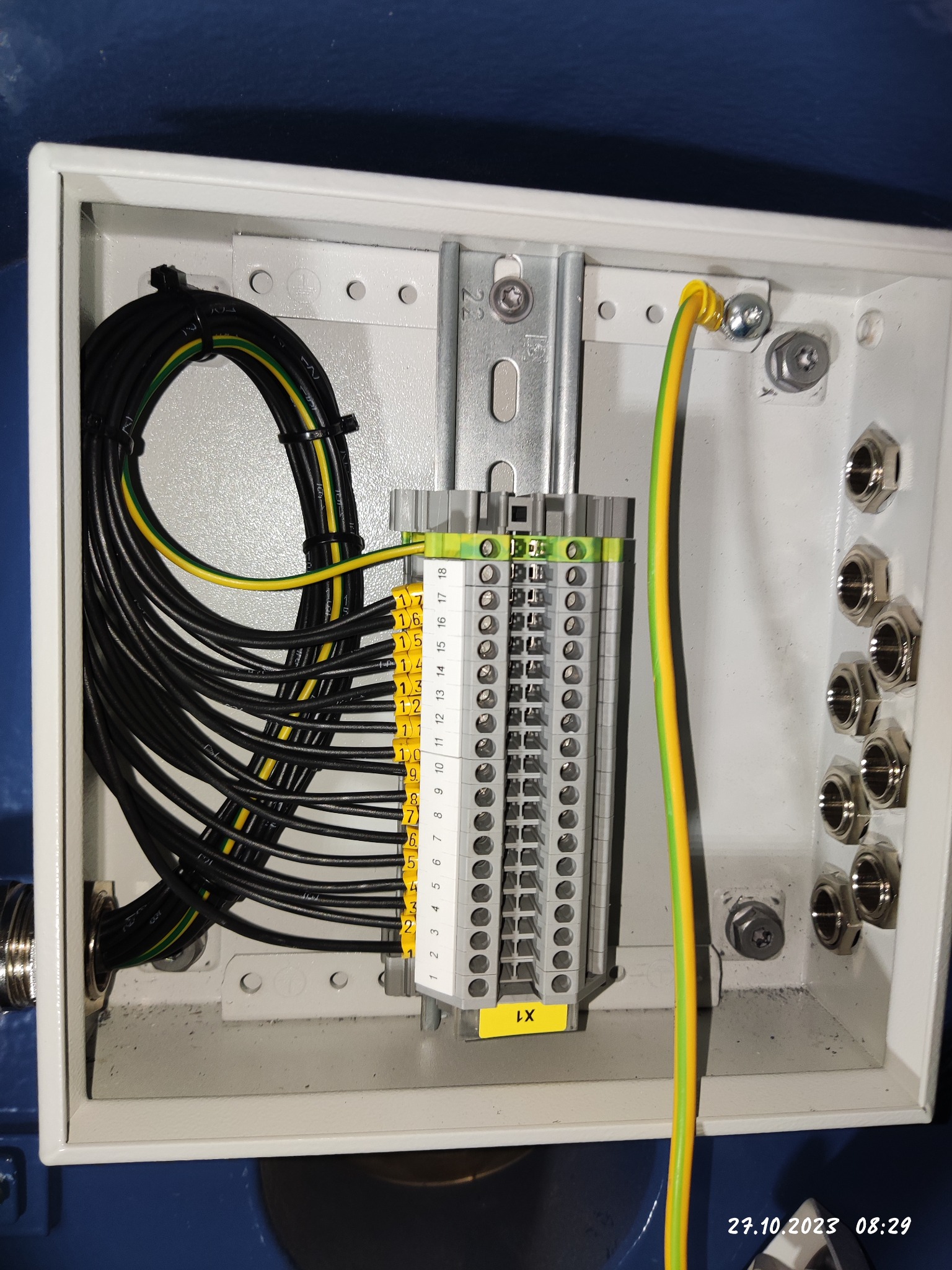 Wnętrze białej skrzynki elektrycznej z zamontowaną listwą zaciskową, podłączonymi przewodami czarnymi i żółto-zielonymi, widoczne naklejki z numeracją.