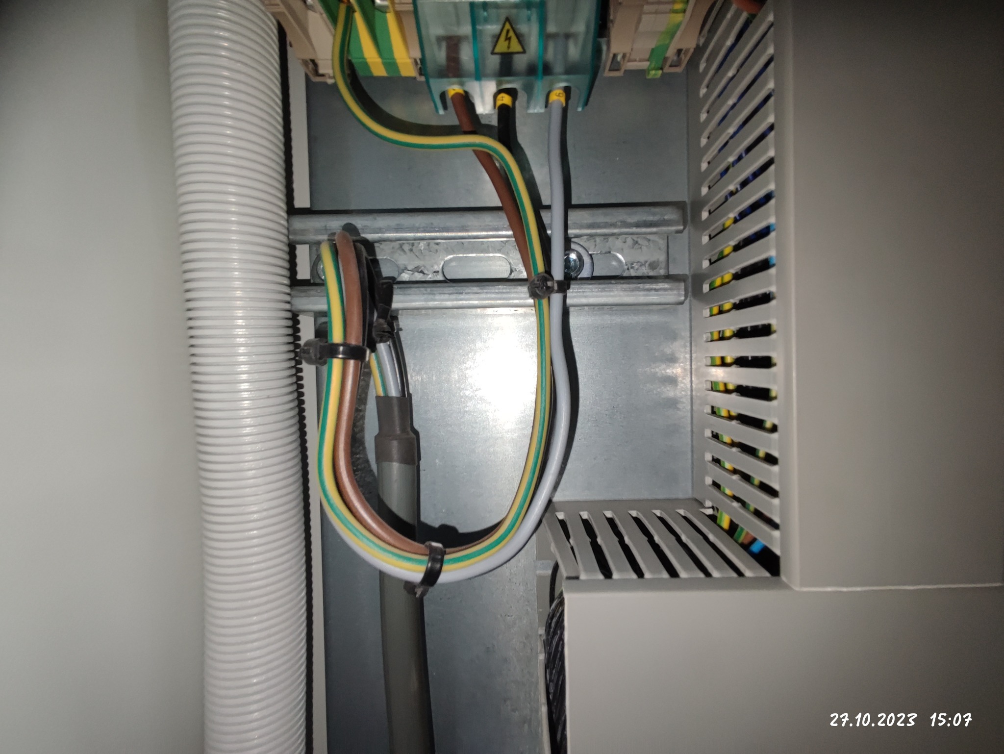 Wewnętrzny widok szafy elektrycznej z przewodami oznaczonymi kolorami, zamocowanymi na szynie DIN i w peszlu ochronnym, widoczny symbol ostrzegawczy wysokiego napięcia na listwie zaciskowej.