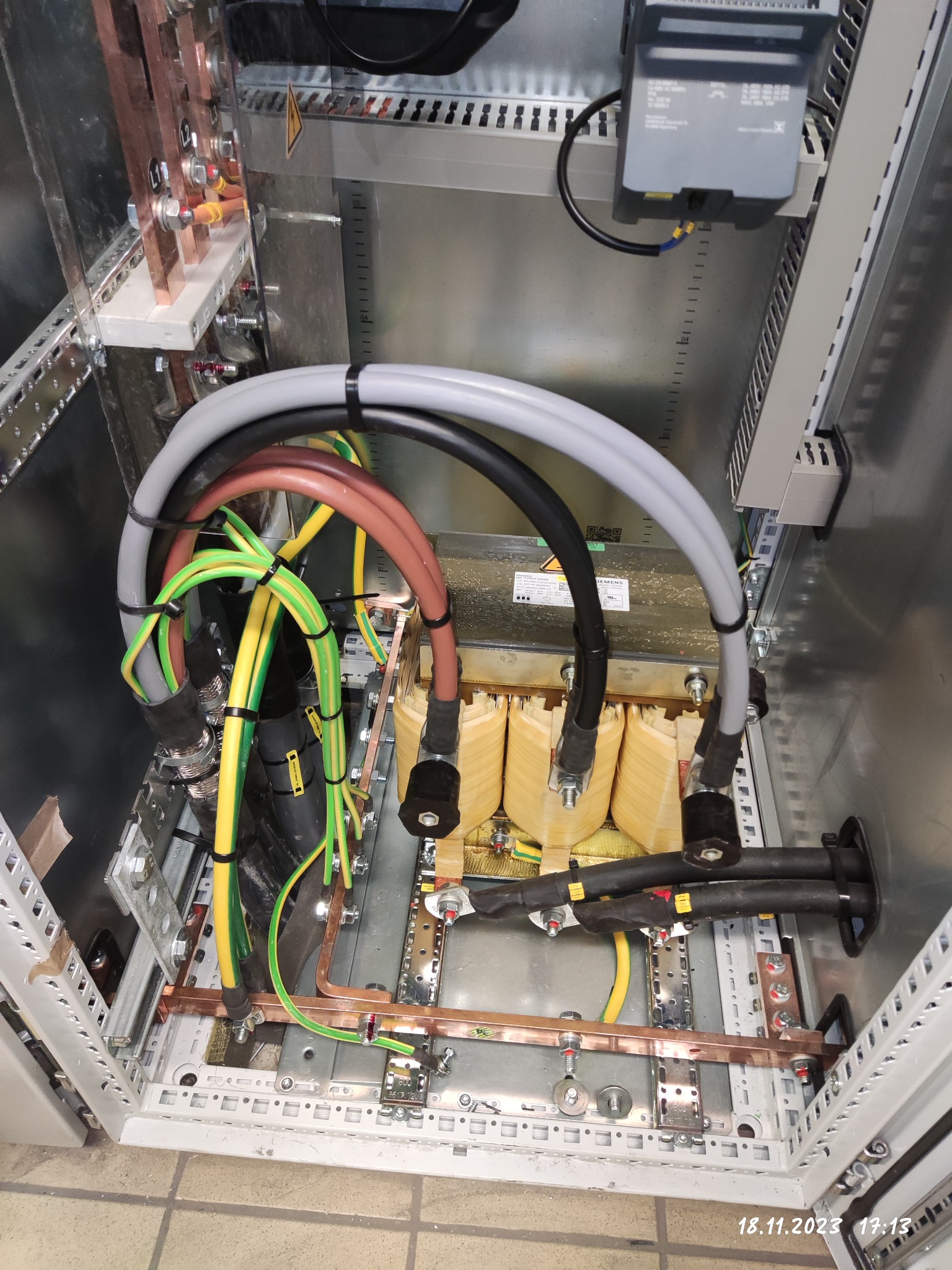 Wnętrze szafy elektrycznej z transformatorem trójfazowym, widoczne przewody o różnych kolorach i przekrojach, szyny miedziane oraz elementy montażowe, całość w metalowej obudowie.