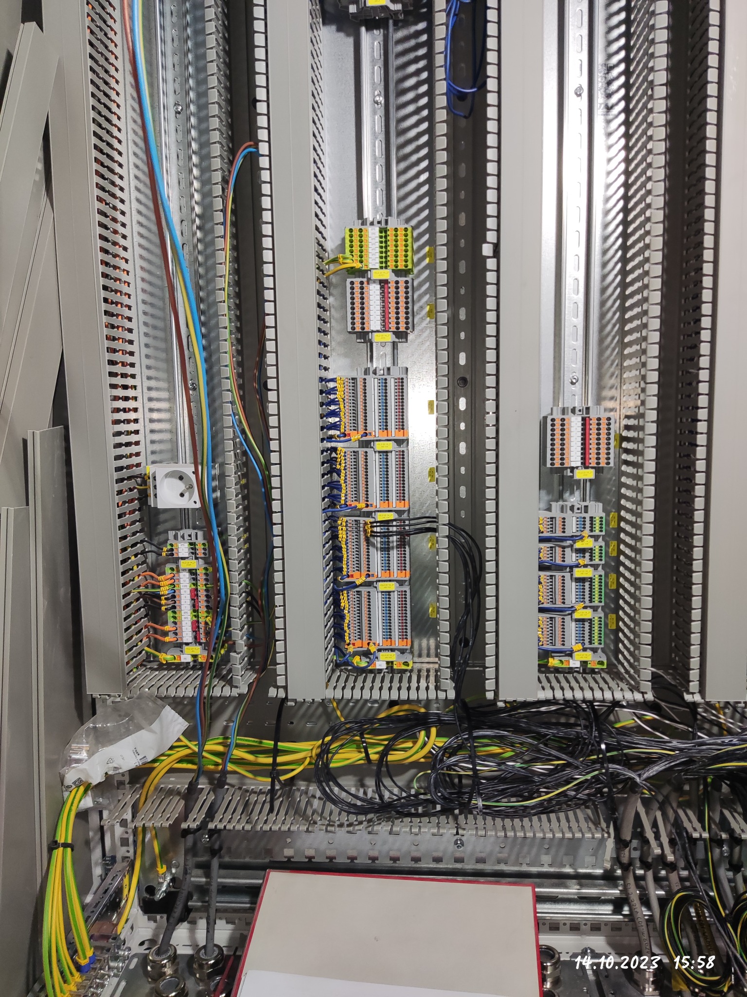 Rozdzielnica elektryczna w trakcie montażu, widoczne liczne przewody, złączki i szyny montażowe. Część przewodów ułożona schludnie, pozostałe w nieładzie. Widoczne gniazdo elektryczne.