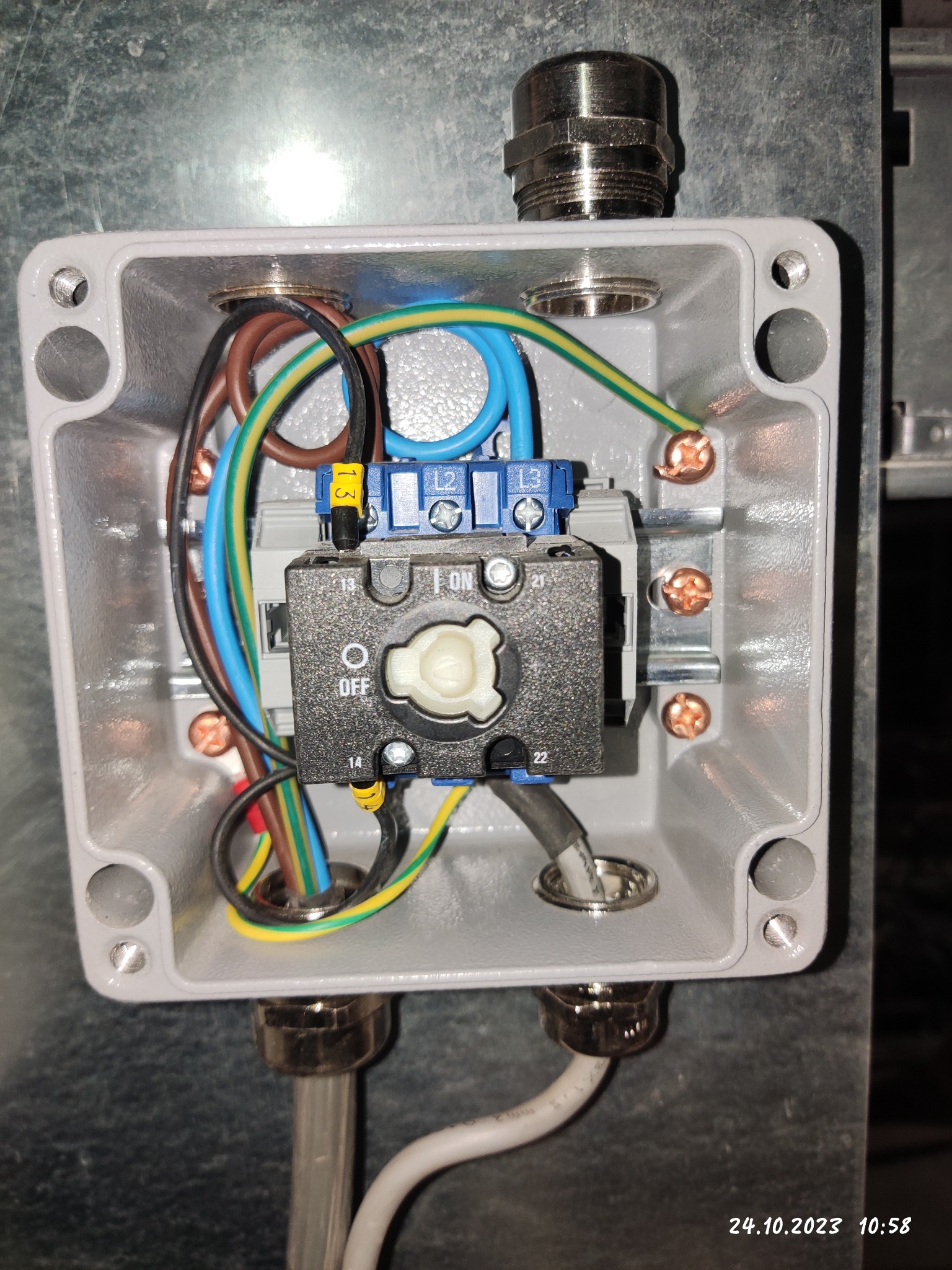 Szara puszka instalacyjna z przełącznikiem obrotowym w pozycji OFF, widoczne przewody z oznaczeniami, zamontowana na szarej powierzchni.