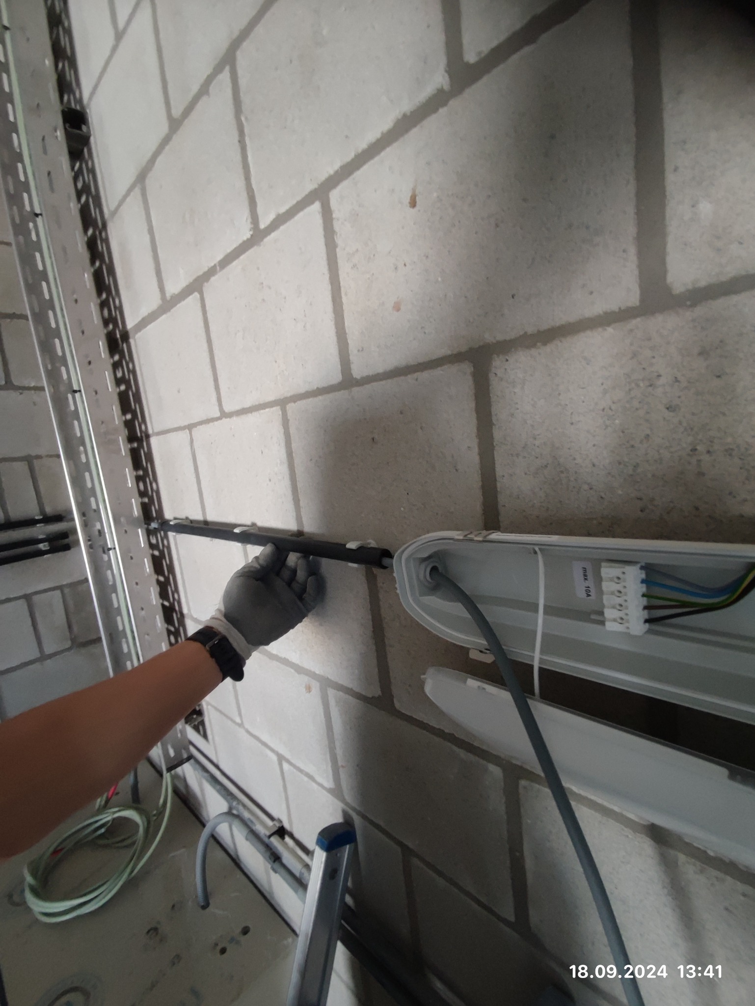 Montaż przewodu elektrycznego w peszlu do puszki oświetleniowej na ścianie z bloczków betonowych. Widoczna drabina i koryto kablowe.