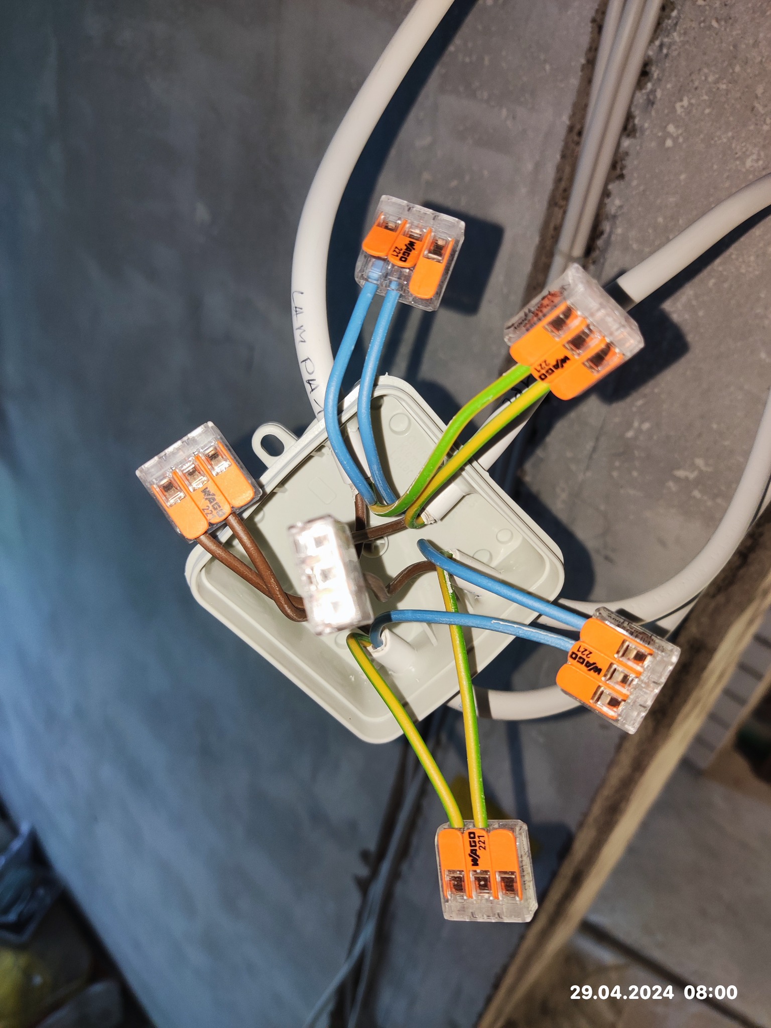 Widok z góry na puszkę elektryczną z podłączonymi przewodami za pomocą szybkozłączek WAGO 221, widoczne przewody w kolorach niebieskim, brązowym i żółto-zielonym.