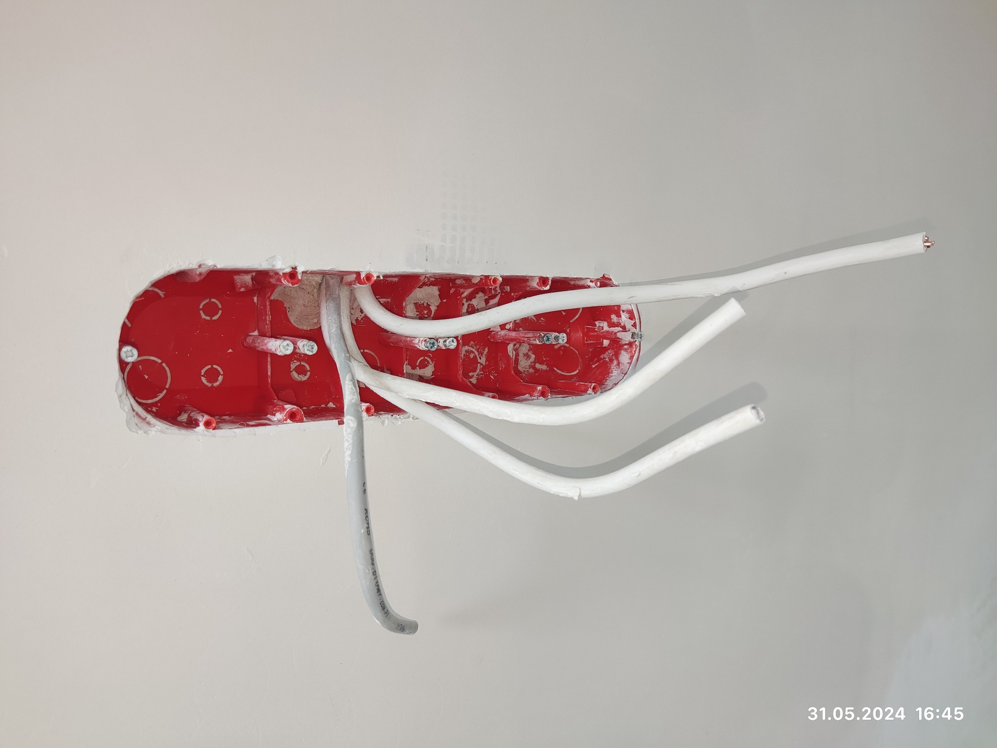 Czerwona puszka elektryczna podtynkowa z wystającymi przewodami w białej ścianie, widoczne ślady gipsu.