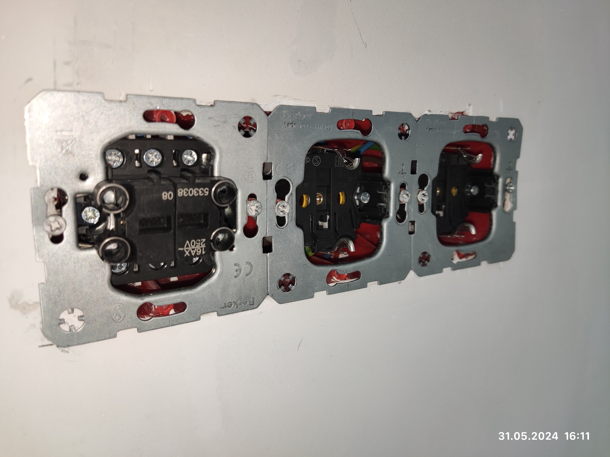 Potrójne gniazdko elektryczne Berker, w trakcie montażu w ścianie, widoczne przewody i mechanizmy wewnętrzne, surowa ściana w tle.