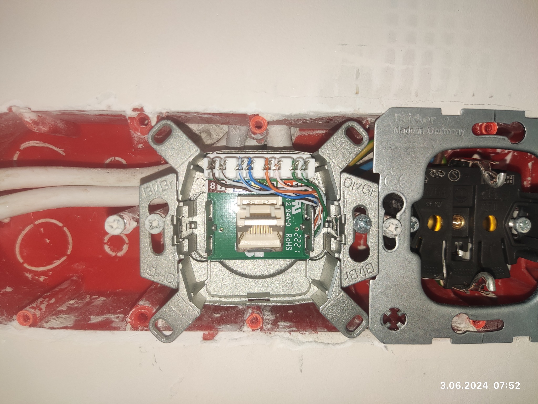 Widok z bliska na gniazdo sieciowe z podłączonymi przewodami oraz gniazdko elektryczne zamontowane w czerwonej puszce instalacyjnej w ścianie, z widocznymi śladami gipsu.