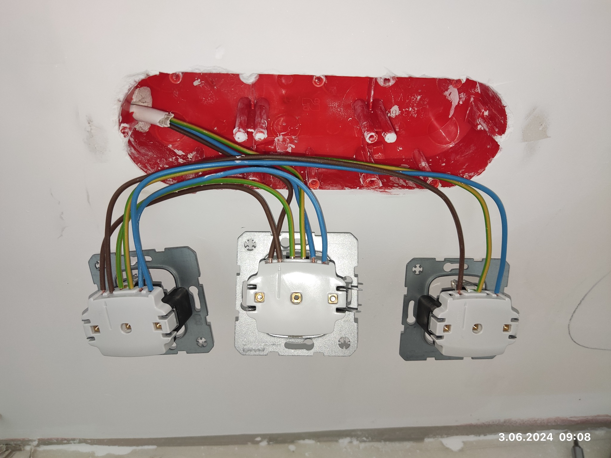 Widok z bliska na podłączenie przewodów do trzech gniazdek elektrycznych w ścianie, widoczna czerwona puszka instalacyjna z przewodami w kolorach: brązowym, niebieskim i zielono-żółtym.