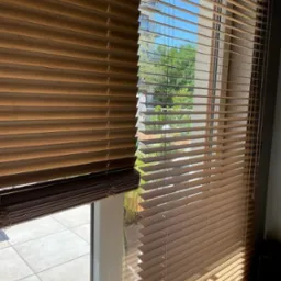 Drewniane żaluzje poziome w odcieniach brązu, częściowo zasłonięte, z widokiem na balkon i zieleń za oknem.