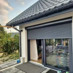 Szara roleta zewnętrzna częściowo opuszczona na oknie tarasowym, widok z zewnątrz na patio z szarą kostką brukową i butami dziecięcymi przy wejściu.