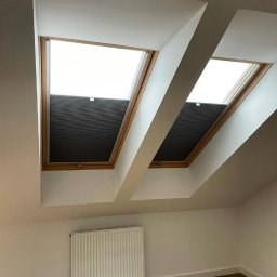 Top Okna Janusz Szewczyk - Dwa okna dachowe z drewnianą ramą, wyposażone w szare, częściowo zasłonięte osłony okienne typu plaster miodu, widoczne w jasnym, białym wnętrzu poddasza z grzejnikiem na ścianie.