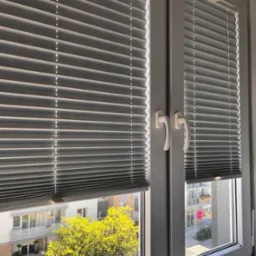 Szare plisy okienne zamontowane na dwuskrzydłowym oknie PCV, widok na budynek mieszkalny i drzewo zza okna.