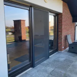 Zewnętrzna moskitiera przesuwna w czarnej ramie, zamontowana na tarasie z widokiem na ogród.