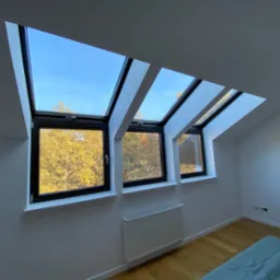 Trzy okna PCV dachowe z czarnymi ramami w białym pokoju na poddaszu, widok na jesienne drzewa i błękitne niebo.