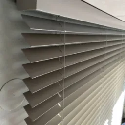 Szare poziome lamele częściowo zasłoniętych żaluzji drewnianych, widoczne sznurki i mechanizm mocujący, z jasnym światłem w tle.