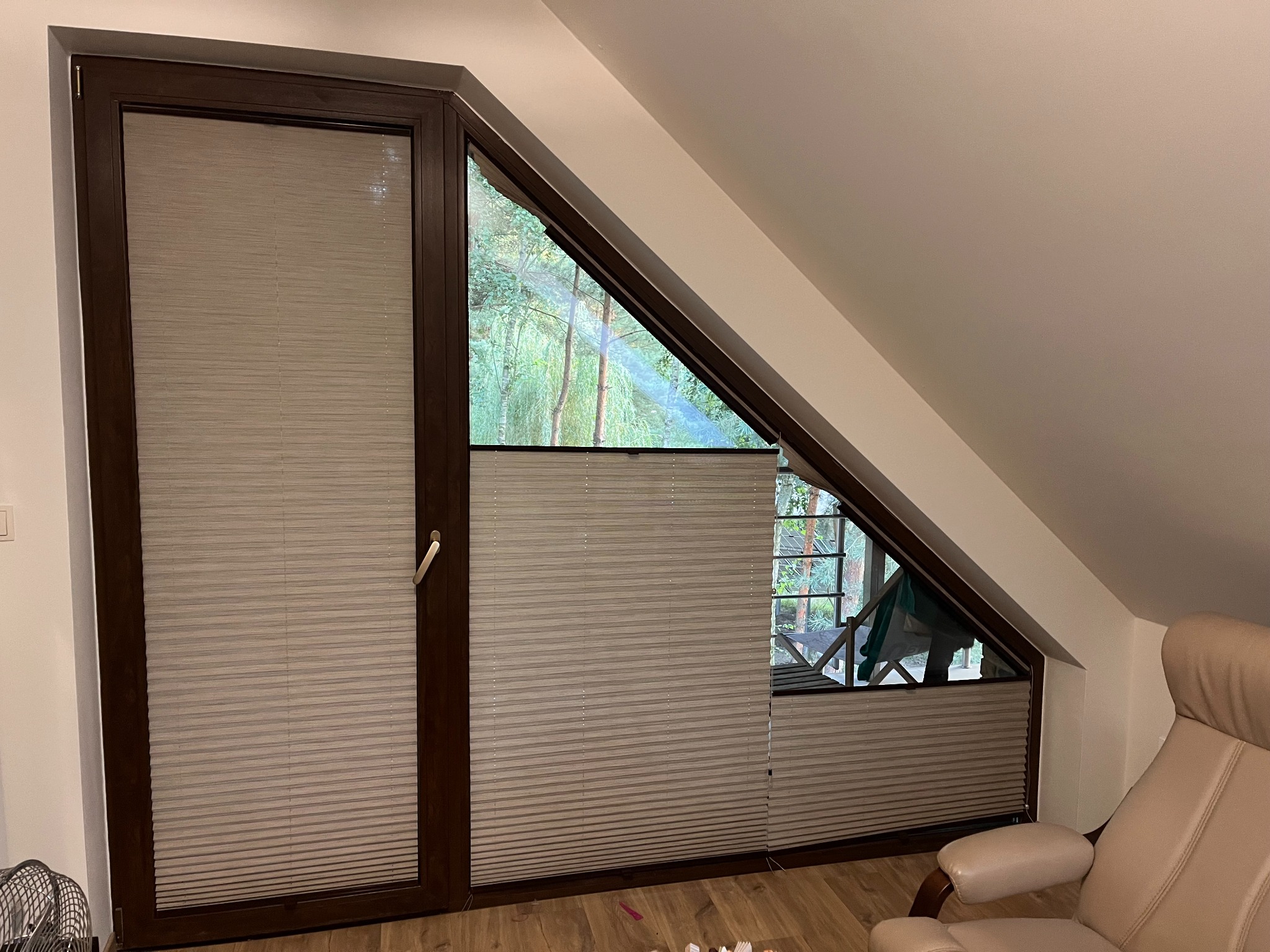 Okno na poddaszu z zamontowanymi plisami w brązowej ramie, widok na drzewa z zewnątrz.