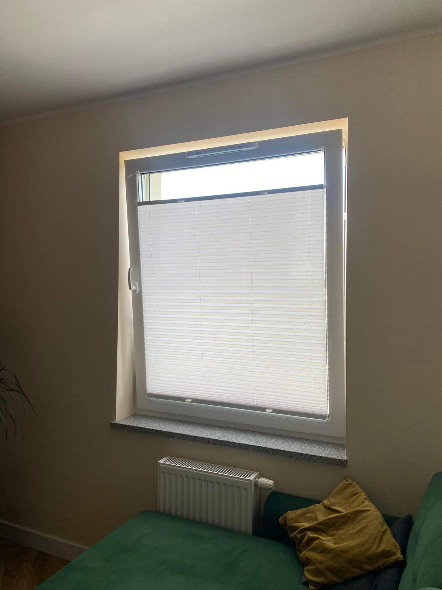 Zamknięta, biała plisa okienna w oknie PCV z szarym parapetem we Wrocławiu, widok z wnętrza pomieszczenia na tle beżowej ściany i fragmentu zielonej kanapy.