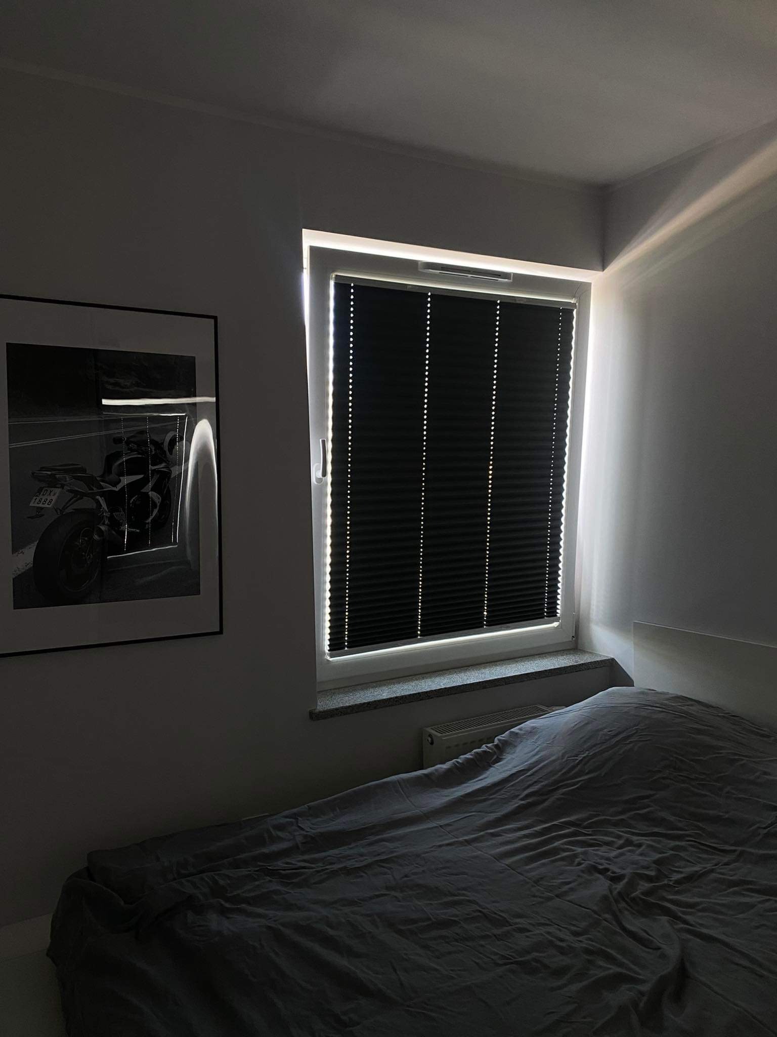 Ciemne plisy okienne zamontowane w oknie sypialni, z widocznym fragmentem łóżka i czarno-białym plakatem motocykla na ścianie, oświetlone popołudniowym słońcem.