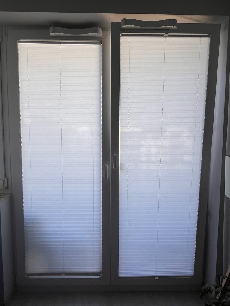 Zamknięte, białe plisy na dwuskrzydłowym oknie PCV, widoczne mechanizmy sterujące