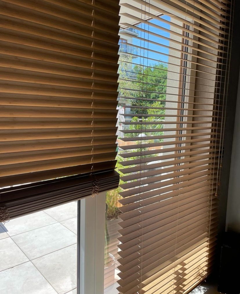 Drewniane żaluzje poziome w odcieniach brązu, częściowo zasłonięte, z widokiem na balkon i zieleń za oknem.