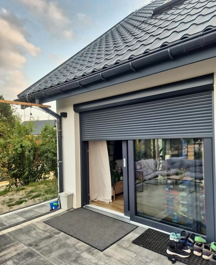 Szara roleta zewnętrzna częściowo opuszczona na oknie tarasowym, widok z zewnątrz na patio z szarą kostką brukową i butami dziecięcymi przy wejściu.