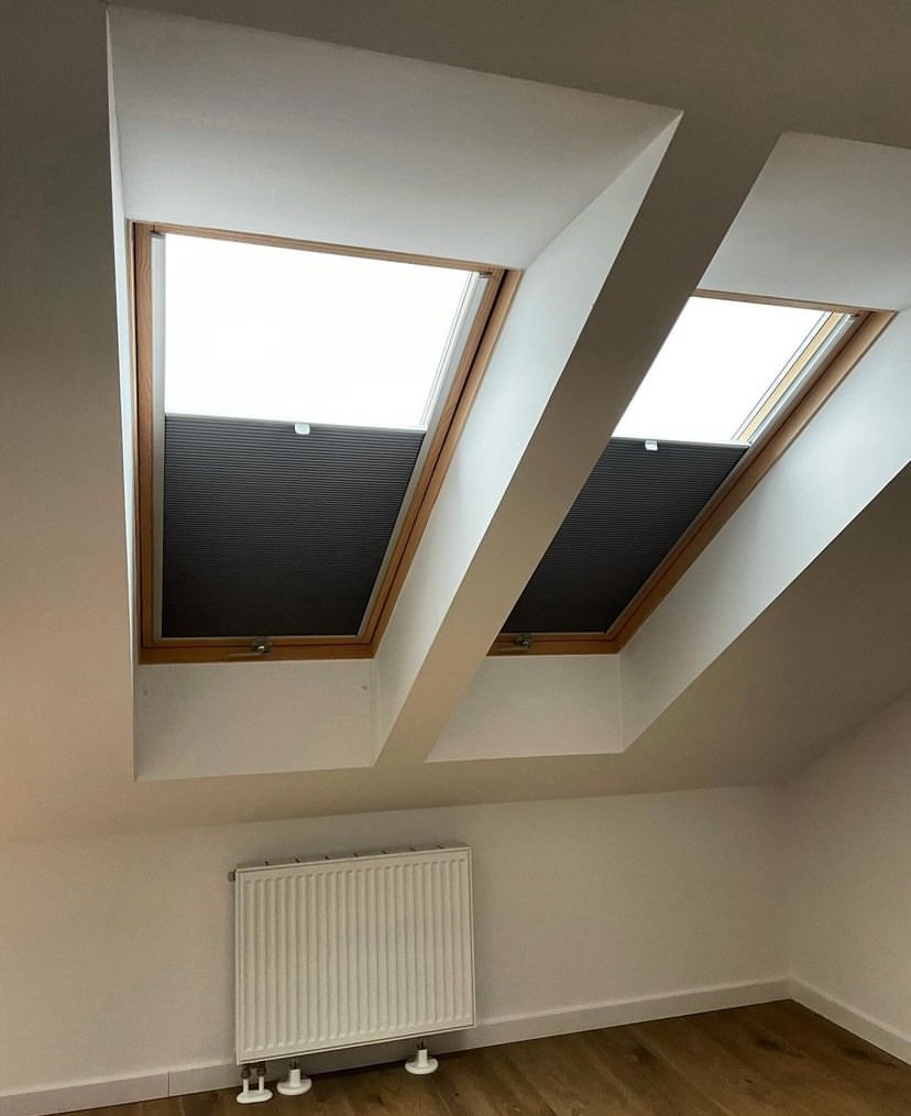 Dwa okna dachowe z drewnianą ramą, wyposażone w szare, częściowo zasłonięte osłony okienne typu plaster miodu, widoczne w jasnym, białym wnętrzu poddasza z grzejnikiem na ścianie.