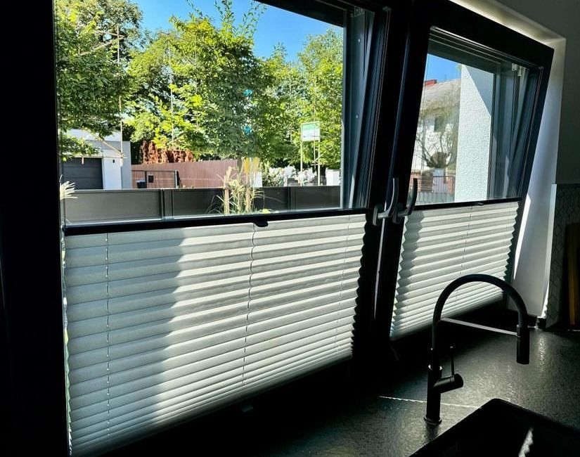 Zamontowane jasne plisy w oknie PCV z widokiem na zieleń ogrodu, widoczny fragment czarnej baterii kuchennej.