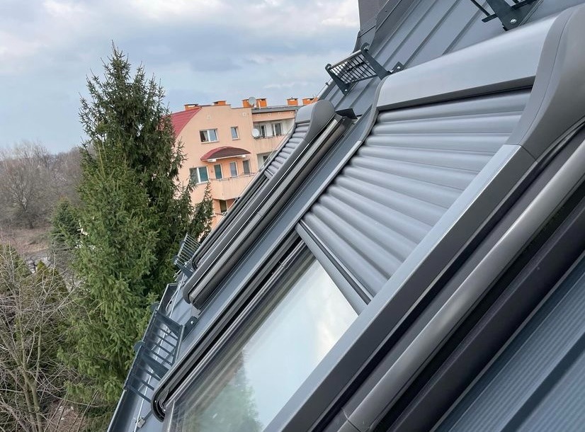 Szare rolety zewnętrzne zamontowane na oknach dachowych, widok z zewnątrz, na tle drzew i budynku mieszkalnego.