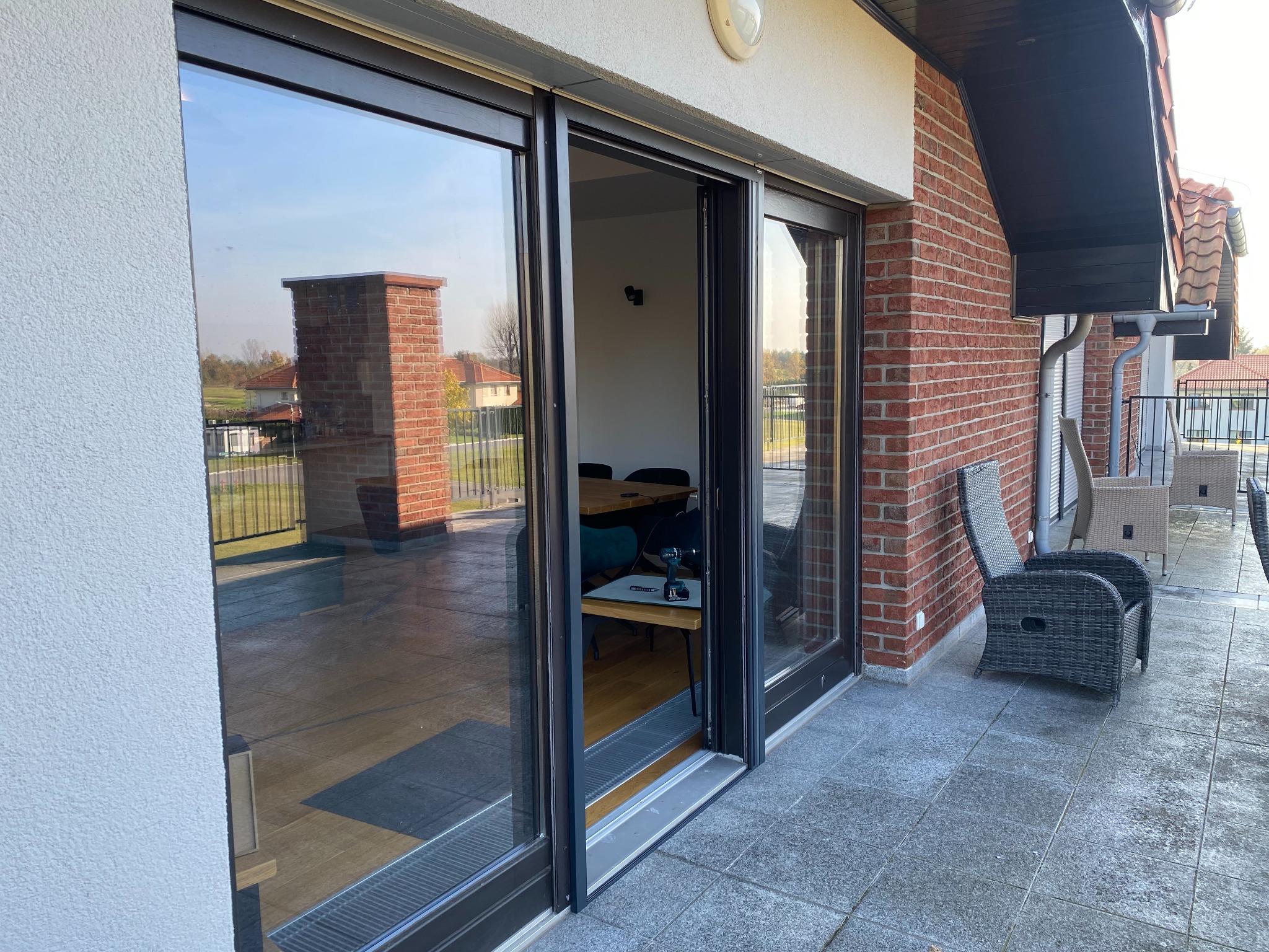 Przesuwne okno tarasowe z czarnymi ramami, widok na wnętrze z narzędziem na stole, ceglana elewacja i meble ogrodowe na zewnątrz.