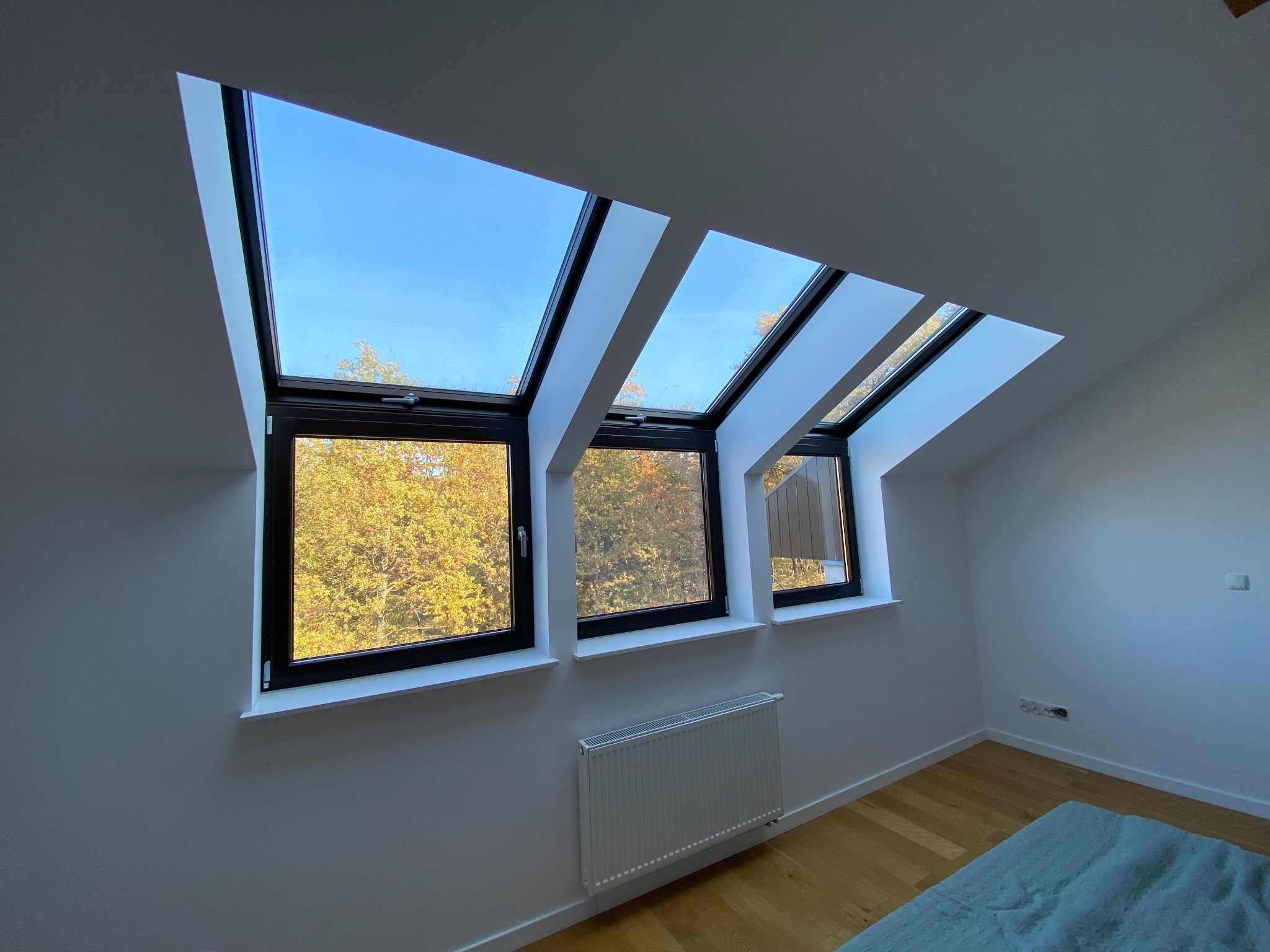 Trzy okna PCV dachowe z czarnymi ramami w białym pokoju na poddaszu, widok na jesienne drzewa i błękitne niebo.