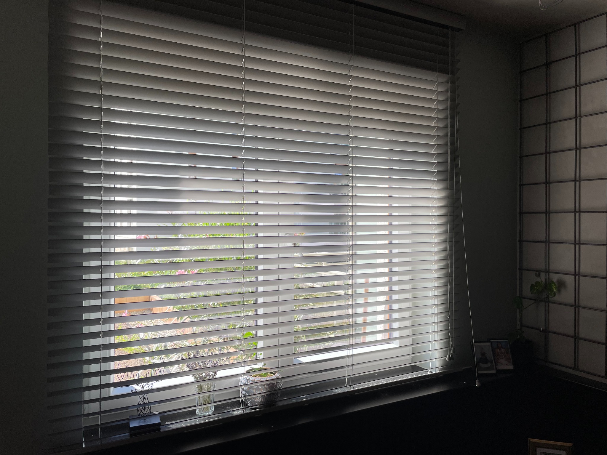 Poziome, białe listwy osłaniają okno, widok na ogród z zewnątrz, dekoracje na parapecie.