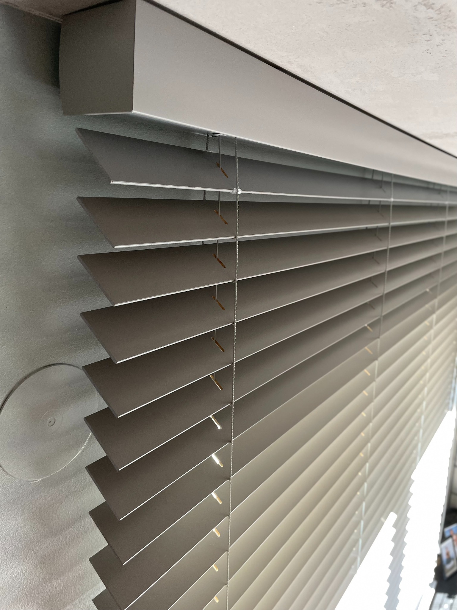 Szare poziome lamele częściowo zasłoniętych żaluzji drewnianych, widoczne sznurki i mechanizm mocujący, z jasnym światłem w tle.