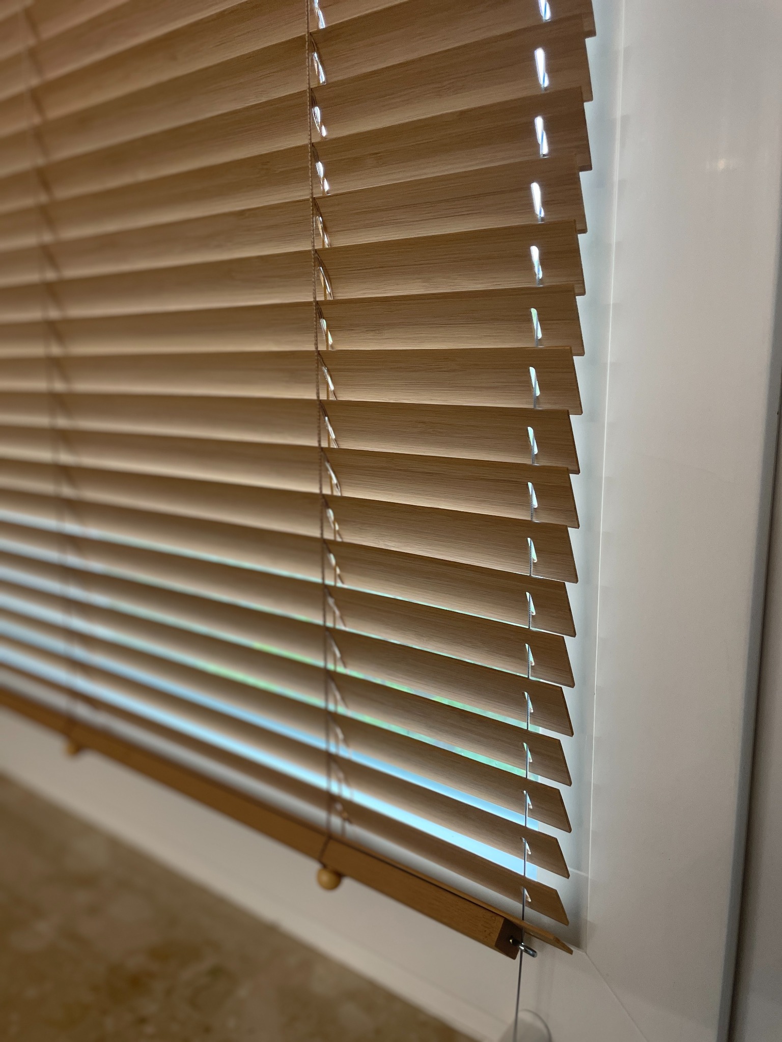 Jasne, drewniane żaluzje poziome zamontowane w białej ramie okiennej, widok z bliska, delikatne światło dzienne prześwitujące między lamelami.