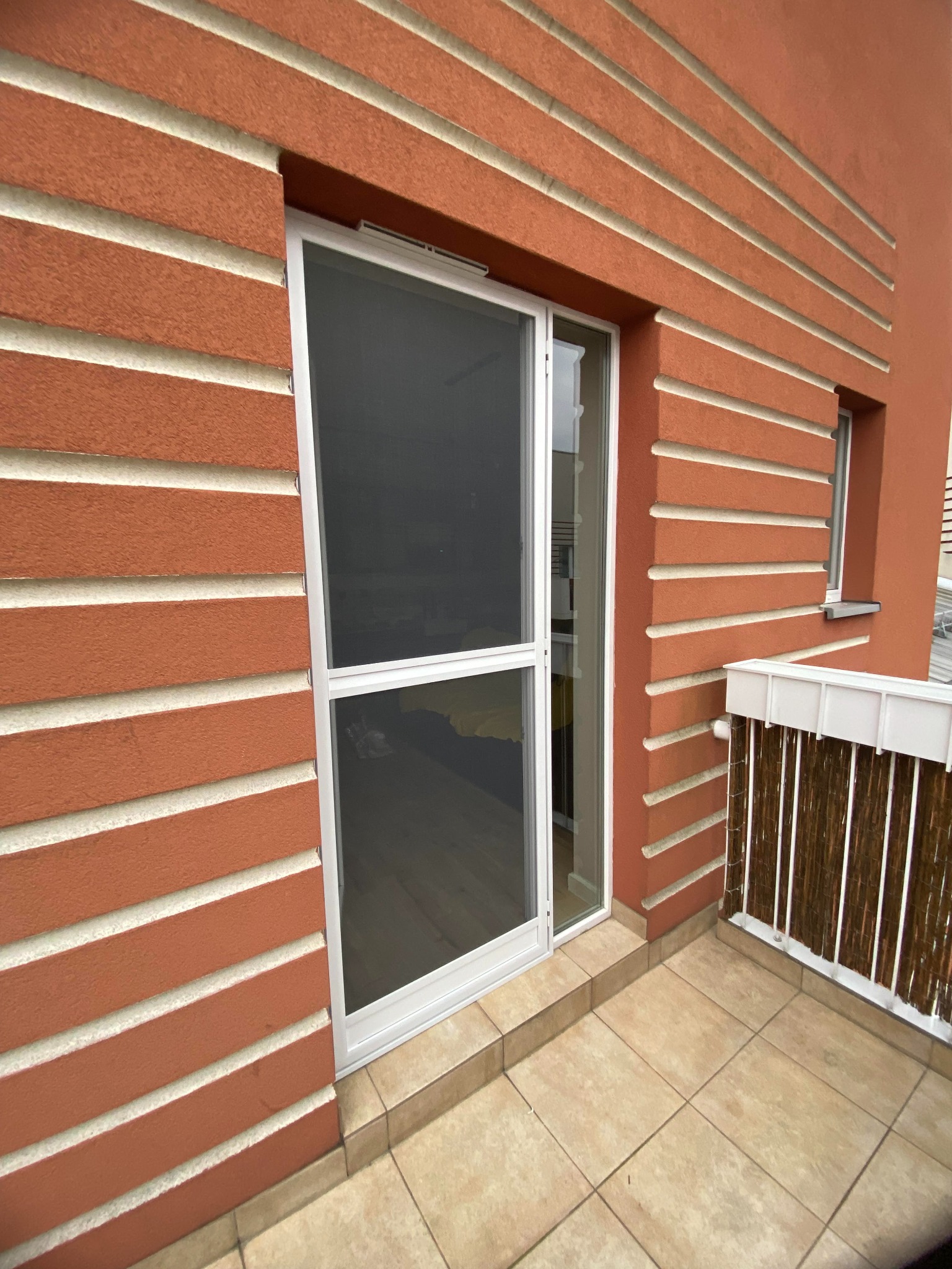 Balkon z pomarańczową elewacją i białymi listwami, białe okno przesuwne z moskitierą.